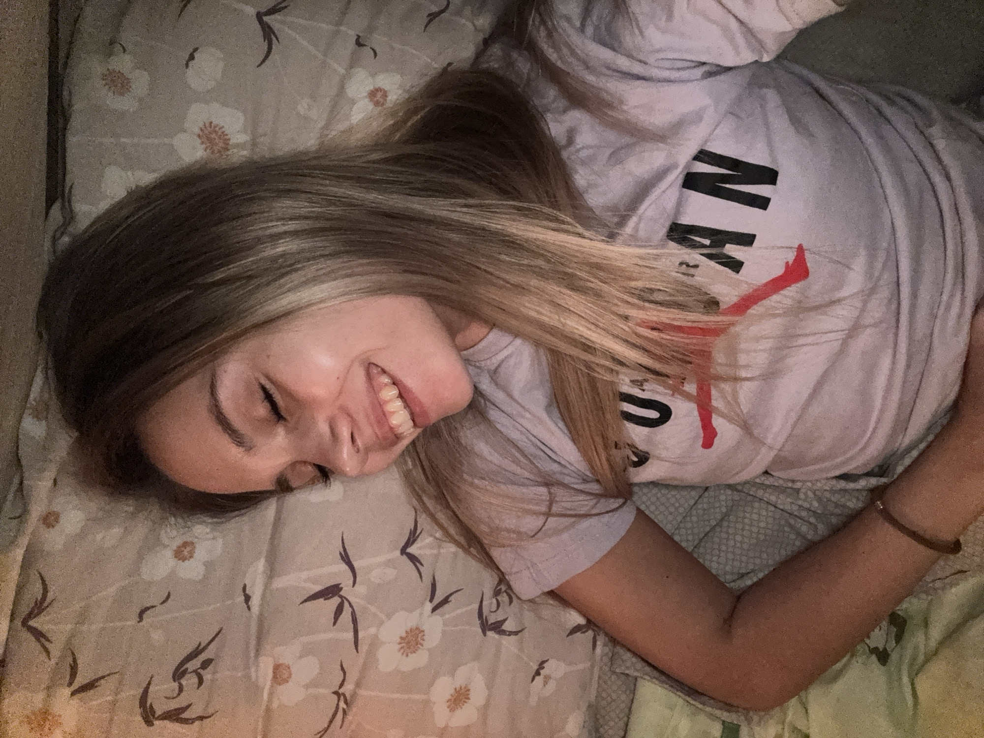 A young woman with blonde hair and closed eyes smiles in bed. She is wearing a light purple t-shirt with the word "JORDAN" printed on it in black and red. The background is a bed with a floral patterned duvet. There are no identifiable landmarks or information that would place this image in Chaikovsky, Russia.
