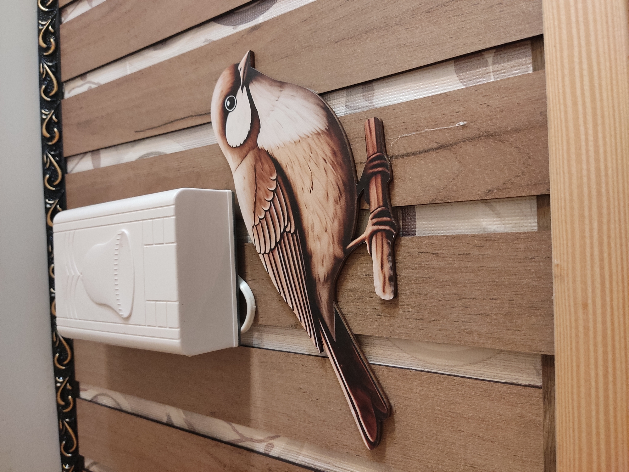 The image shows a decorative bird cutout on a wooden slatted surface, with a white doorbell or chime box in the background. The bird is depicted as a brown and white sparrow perched on a thin branch. The doorbell is rectangular with a raised bell shape on its front. The background consists of wooden slats with a textured fabric-like material visible between them. The image is taken from a high angle, looking down on the objects. The scene appears to be indoors, likely in a residential setting. There is no indication of the city of Noginsk, Russia, or any specific location.