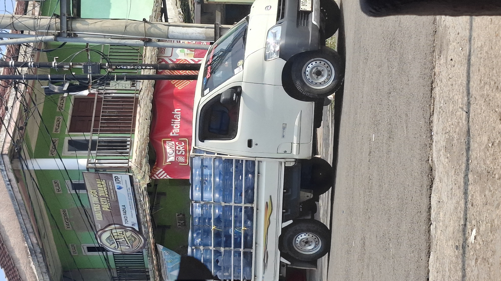 A white pickup truck is parked on the side of a road in Cilaku, Indonesia. The truck is loaded with numerous blue water jugs. A building with green and white paint is visible in the background, with a balcony on the second floor. A red banner with white text is hanging from the building. Utility poles with a dense network of wires are also present. The road surface is asphalt.