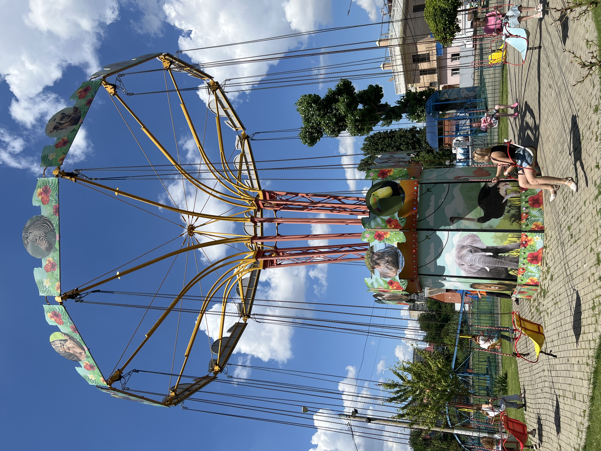 In Krasnodar, Russia, a swing ride at an amusement park is depicted on a sunny day with blue skies and scattered clouds. The ride features a central platform decorated with colorful images of animals like elephants, lions, hippos, zebras, and toucans, along with tropical flowers. Suspended from a yellow, multi-armed structure by chains are red and yellow swings, some occupied by children. A young woman is seated in a swing in the foreground, wearing a black top and denim shorts, while another young woman in a pink shirt and denim shorts is in a swing further back. In the background, a playground structure is visible, as well as residential buildings and trees. The ground is paved with interlocking bricks.