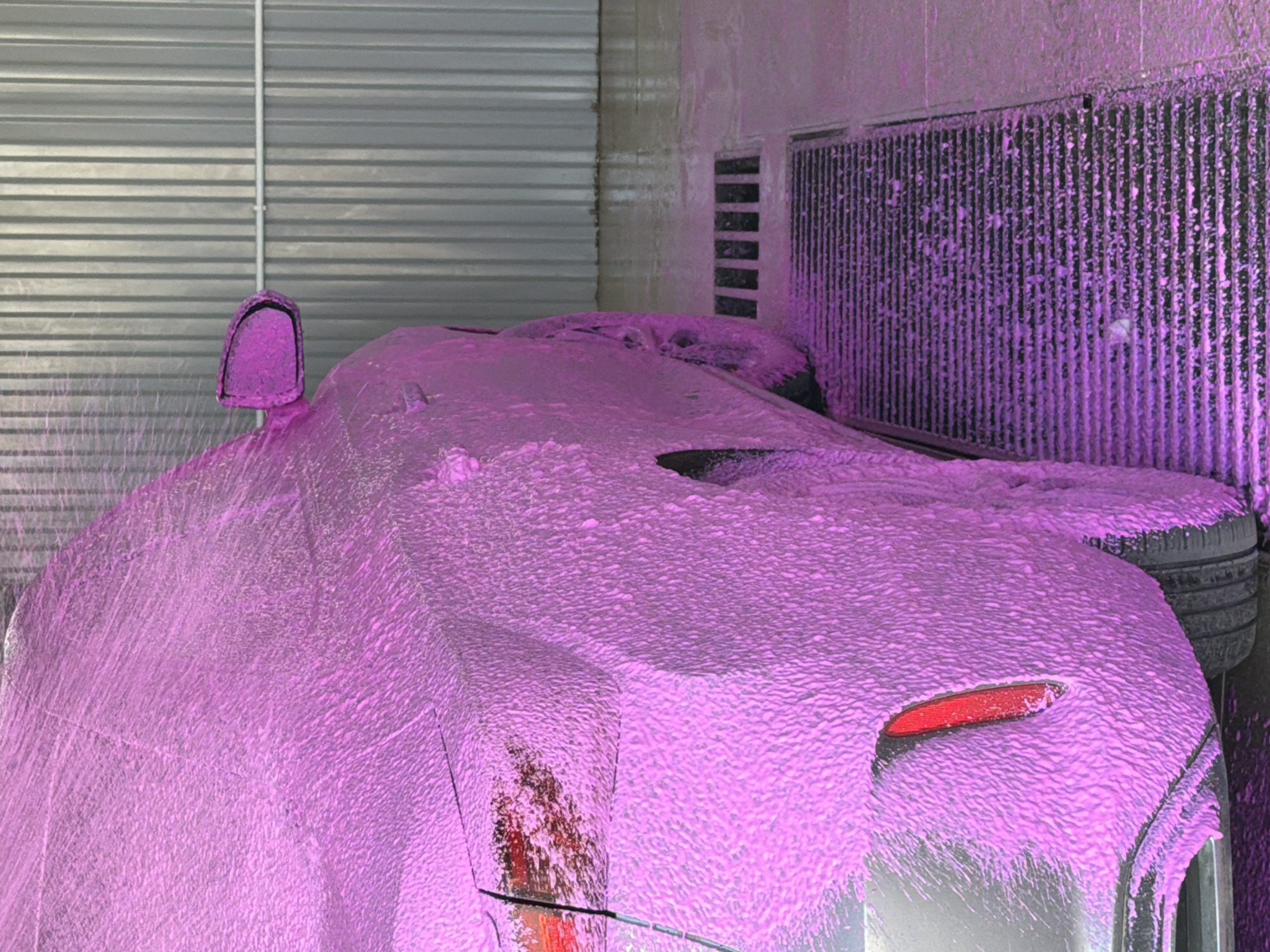 A car covered in pink foam stands inside a car wash facility in Chaikovsky, Russia. The car's taillight and side mirror are partially visible beneath the foam. To the left of the vehicle, there's a corrugated metal wall, and on the right, a metal grating is also covered in pink foam.