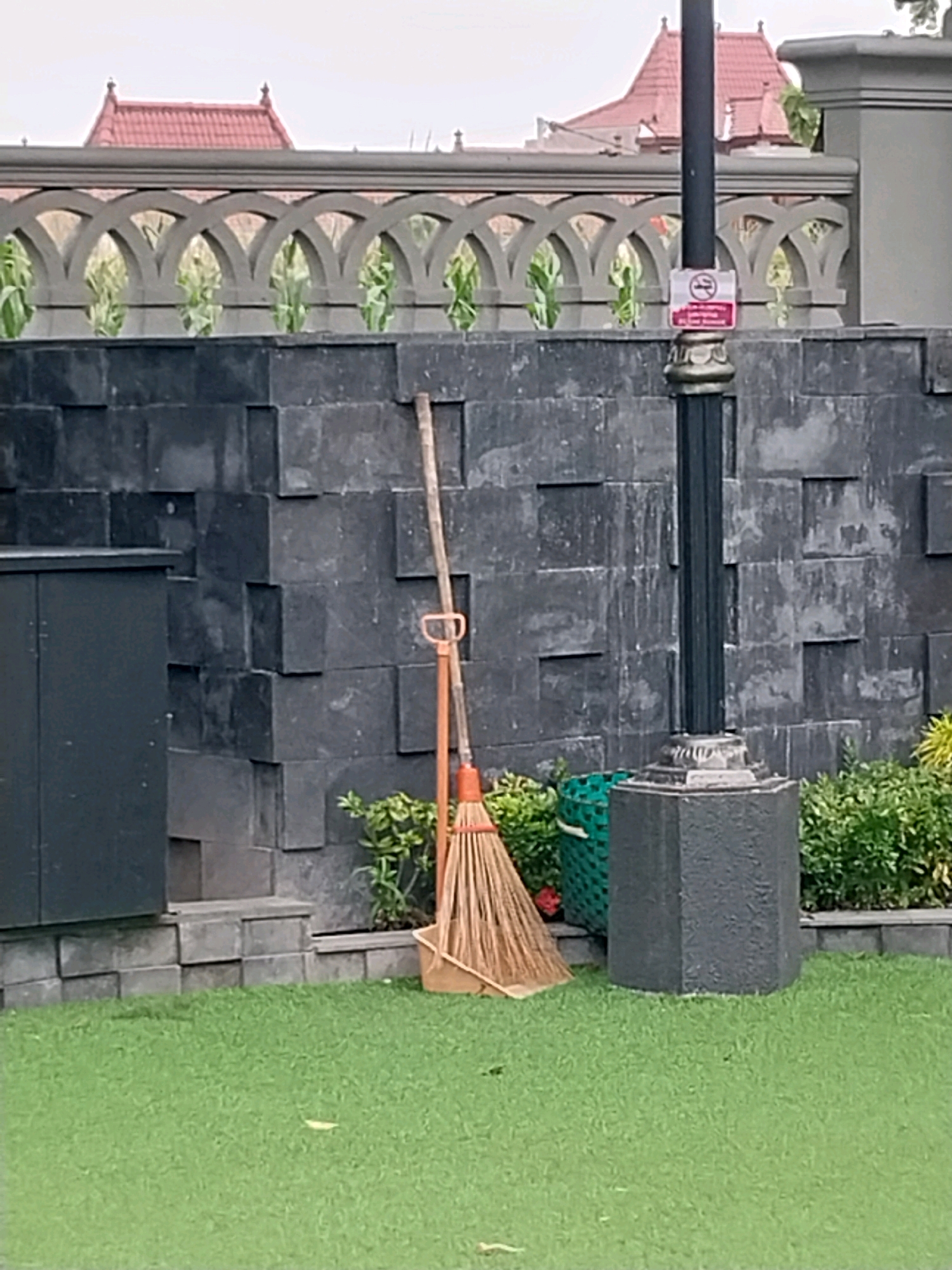 In Bugisan, Indonesia, a broom leans against a wall constructed of dark, rectangular stone blocks. The wall is positioned behind a patch of vibrant green grass. To the left of the broom, a dark gray cabinet is visible. To the right, a black lamppost stands, adorned with a circular sign indicating a prohibition against smoking. Behind the wall, an ornate grey railing with arched designs spans across the frame, and beyond that, a building with a red tile roof is partially visible. Lush green plants grow in front of the wall, and a green woven basket sits near the lamppost.