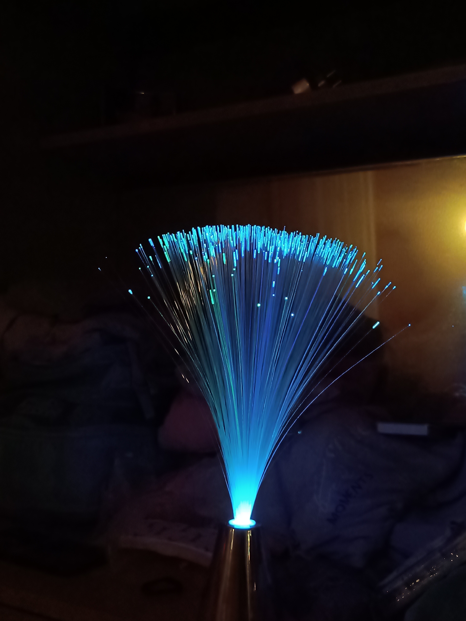 A fiber optic lamp is positioned in a dim room. The lamp has a brass-colored base, with radiating blue, light blue, and green fiber optic strands. The fibers are more concentrated toward the top. In the background, blurred items suggest a room with textiles. The location of Cherkovitsa, Bulgaria, is not discernible from the image.