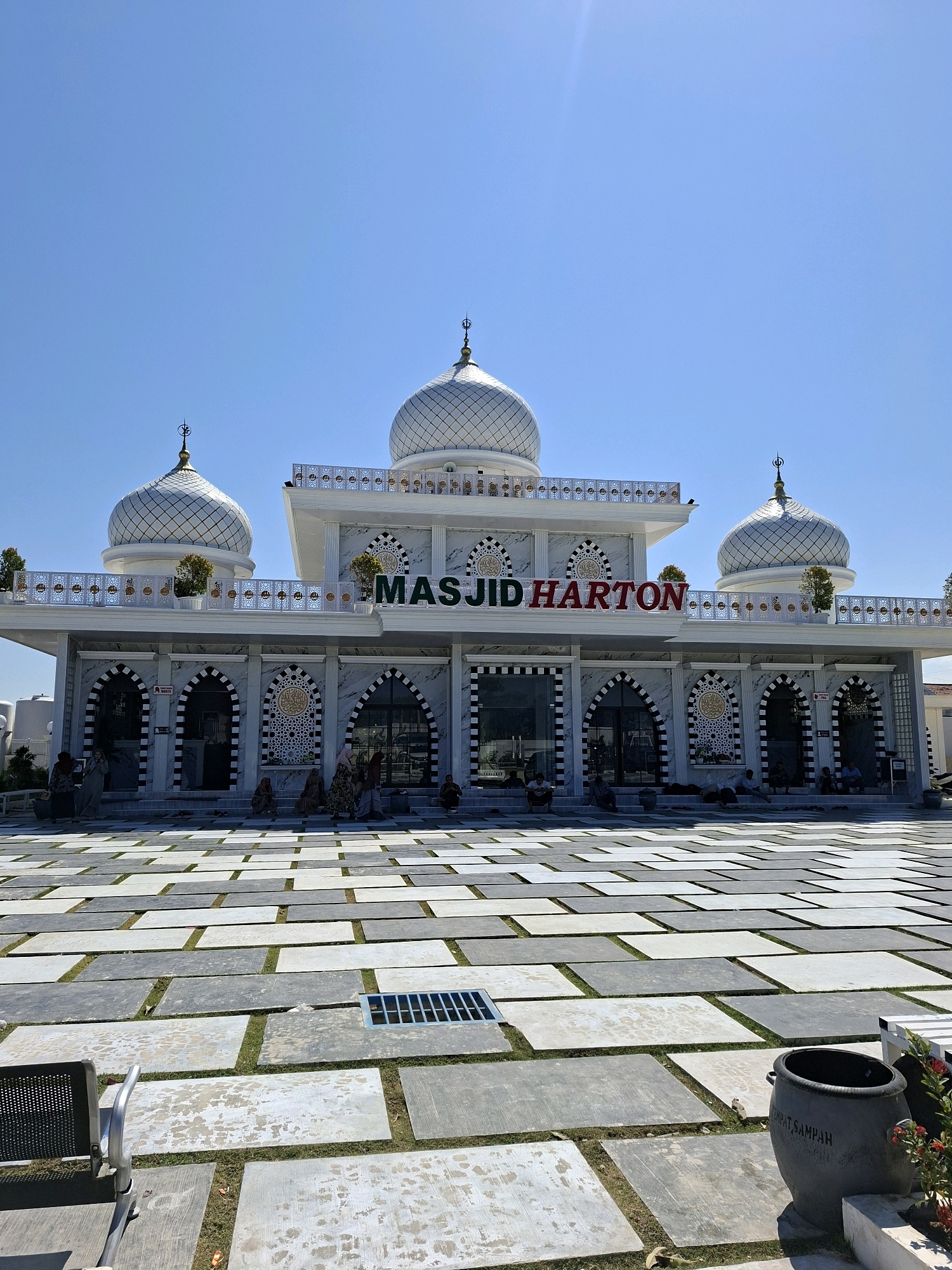 This is a low-angle, full shot of the Masjid Harton in Camplong, Indonesia, under a clear blue sky. The mosque features a white facade with ornate details and multiple silver domes topped with crescent moons. The name "MASJID HARTON" is prominently displayed in red lettering above the entrance. The building has arched doorways and windows, with decorative patterns on the walls. A large paved courtyard in front of the mosque is laid with large, rectangular stone tiles, interspersed with patches of grass. Several people are seated on the steps leading into the mosque, and a few others are standing or walking in the courtyard. In the foreground, a black trash can with Indonesian writing is visible, along with a metal chair. The overall scene depicts a religious site on a bright, sunny day.