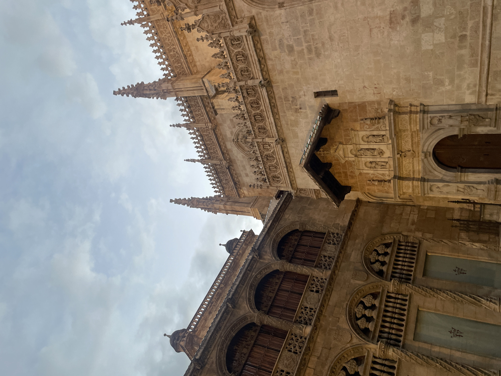 This image depicts the exterior of the Capilla Real (Royal Chapel) in Granada, Spain, viewed from a low angle looking upwards towards a cloudy sky. The architecture is predominantly made of light-colored stone, with intricate details.
On the left, a section of a building features a loggia with three arched openings, each covered by dark wooden shutters. Above these arches, decorative stonework can be seen. The building also has a domed structure at its top left corner, topped with a small spire. Below the loggia, there are two large doors, each adorned with a crest featuring what appears to be intertwined initials and a crown. The stonework around these doors is ornately carved.
To the right, a more elaborate facade of the Royal Chapel is visible. This section is characterized by a series of pointed spires and ornate decorative friezes running along the upper portions of the building. There is a small, pitched roofed structure above an entrance, adorned with sculptures of figures in niches. A large wooden door is visible beneath this structure, set within an arched portal. The walls are constructed from large, rough-hewn stone blocks.
The overall lighting suggests it is either early morning or late afternoon, with the sky overcast and the stone buildings illuminated by diffused light. The scene conveys a sense of historical grandeur and architectural complexity.