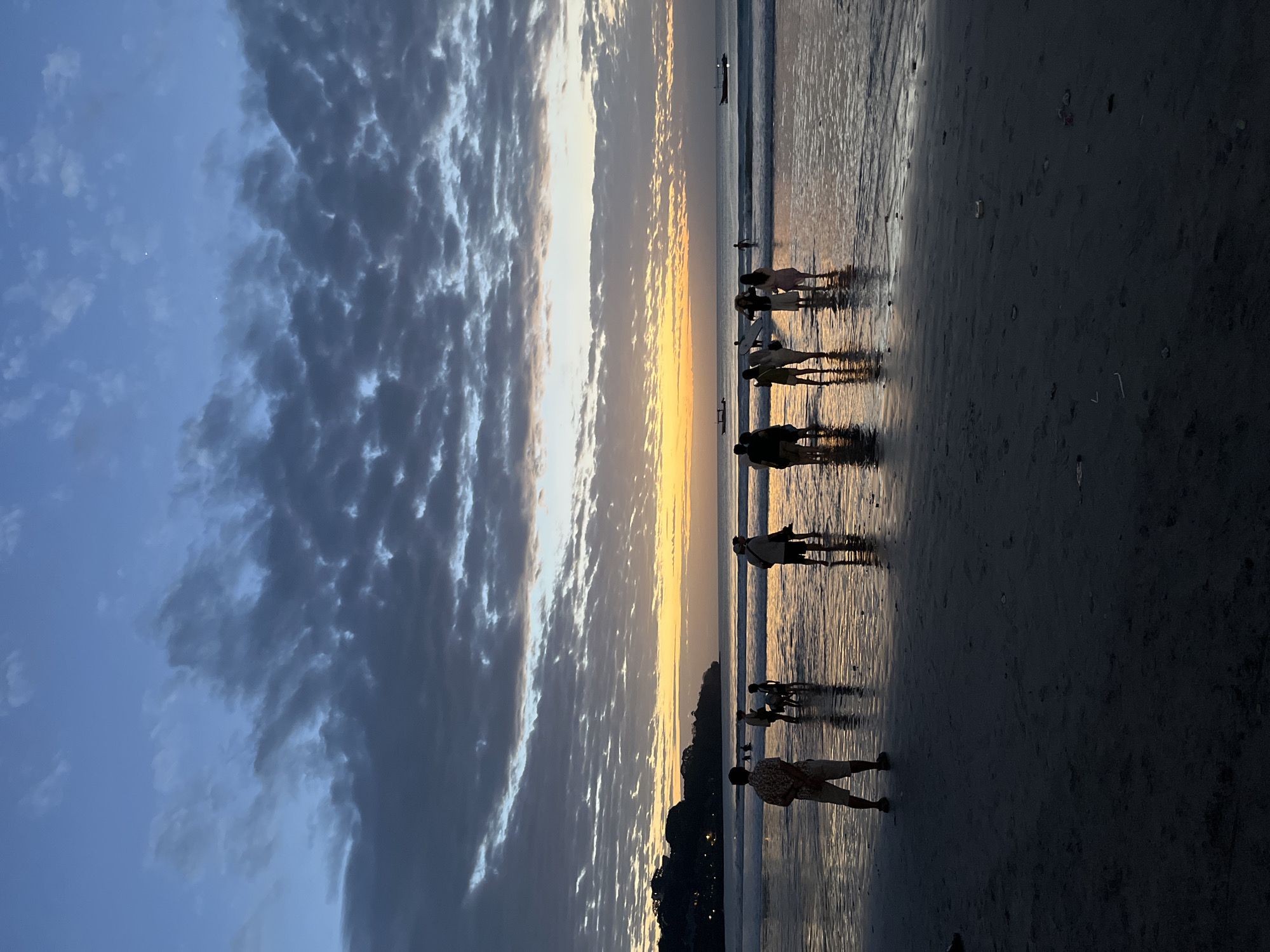The image depicts a serene beach scene at dusk in Kuta Selatan, Indonesia. The sky is a gradient of deep blue at the top, transitioning to lighter blues and oranges near the horizon, painted with dramatic clouds. A warm, golden light emanates from the setting sun, casting a warm glow across the water and wet sand. Several individuals and groups of people are scattered across the beach, silhouetted against the bright sky and reflecting in the shallow water. They are standing, walking, and interacting, their figures creating dark shapes against the vibrant backdrop. The ocean is calm with gentle waves lapping at the shore. In the distance, the outline of a tree-covered hill is visible on the left, with a few scattered lights indicating some settlement. Two small boats are present on the water, adding to the peaceful atmosphere. The foreground of the image is dominated by the dark, wet sand of the beach, reflecting the sky's colors and the silhouettes of the people.