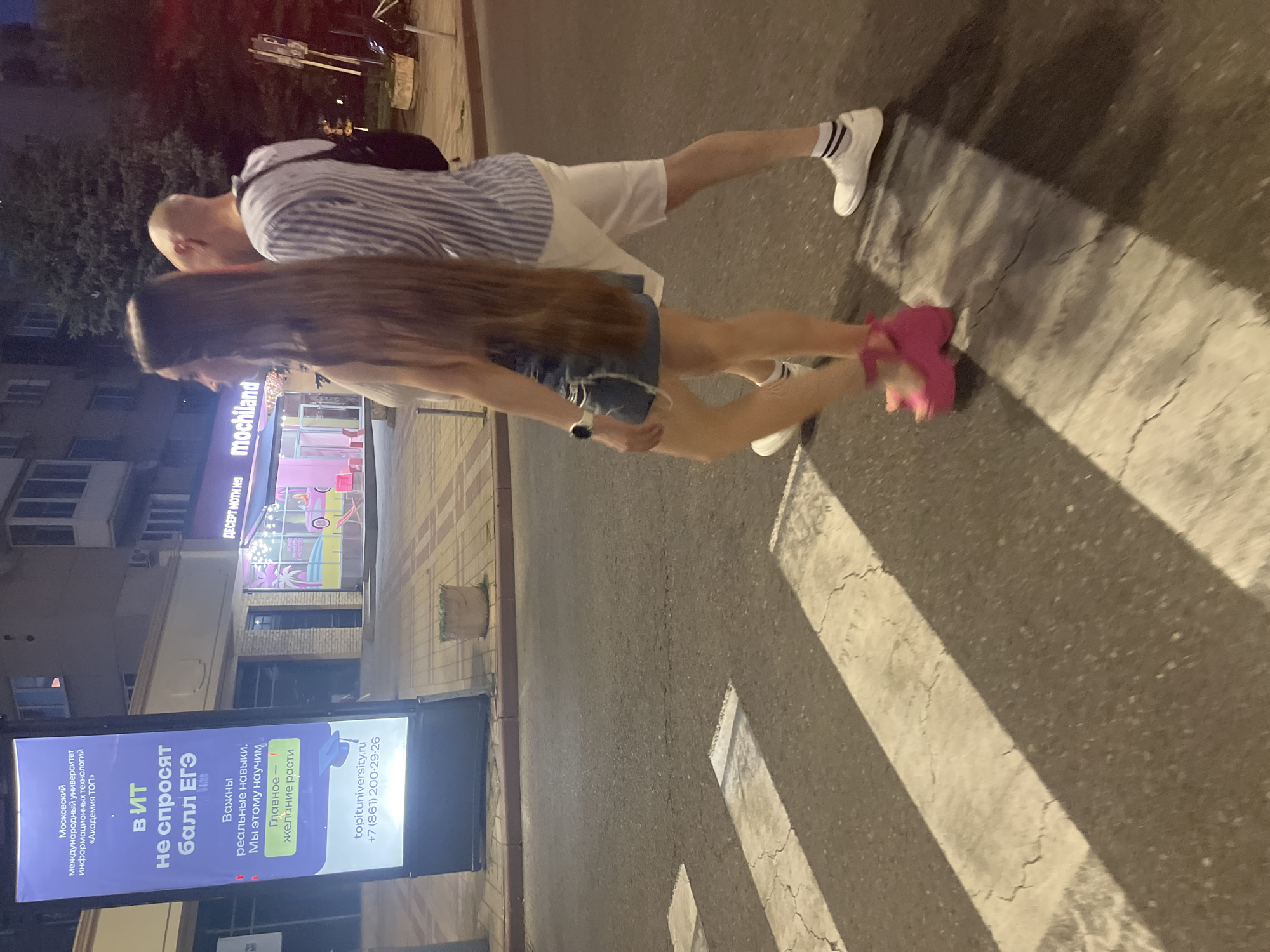 The image shows a street scene at night in Krasnodar, Russia, likely near a commercial area. A young woman with very long brown hair and a denim skirt is walking across a pedestrian crossing, wearing bright pink platform heels. She is accompanied by a man in a striped shirt and white shorts. Behind them, a large illuminated billboard advertises a university, "Moskovskiy mezhdunarodnyy universitet informatsionnykh tekhnologiy 'Akademiya TOP'". The billboard text translates to: "In IT, exam scores are not asked for. What's important are real skills. We teach this. The main thing is the desire to grow." Below this, contact information is provided: "topituniversity.ru" and "+7 (861) 200-29-26". To the right of the billboard, a storefront with neon lighting and the name "mochiland" is visible, suggesting a dessert or ice cream shop. The street is asphalt, and the pedestrian crossing is marked with white stripes. The lighting suggests it is evening or night.