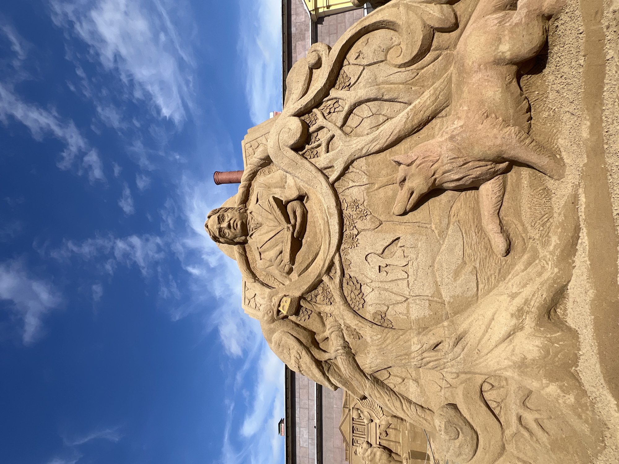This media file features a large, intricately carved sand sculpture, likely a temporary art installation, set against a bright blue sky with wispy white clouds. The sculpture is oriented vertically, showcasing considerable height and detail.
The central figure of the sand art is a bust of a man, depicted at the upper part of the relief, holding what appears to be an open book or similar object. His expression is contemplative. Surrounding the man and integrating into the overall design are elaborate carvings of nature, including prominent, gnarled tree branches and detailed foliage with leaves. To the right of the man, a well-defined figure of a fox or wolf-like animal is carved, appearing to be in motion within the sandy landscape. Further down, other smaller figures, possibly a bird or small animal, are subtly incorporated into the natural scenery. The lower section of the sculpture hints at architectural elements, with carvings that resemble small buildings or houses, possibly representing a village or town.
The entire sculpture exhibits remarkable craftsmanship in sand, with varying textures and depths creating a dynamic and engaging relief. The clear blue sky with scattered clouds suggests a bright, sunny daytime setting. In the background, glimpses of buildings with yellow, grey, and reddish-brown elements are visible, along with a reddish-brown cylindrical object, possibly a chimney, protruding against the sky. No visible text is present in the image. Given the location context, this artistic display is situated in Saint Petersburg, Russia.