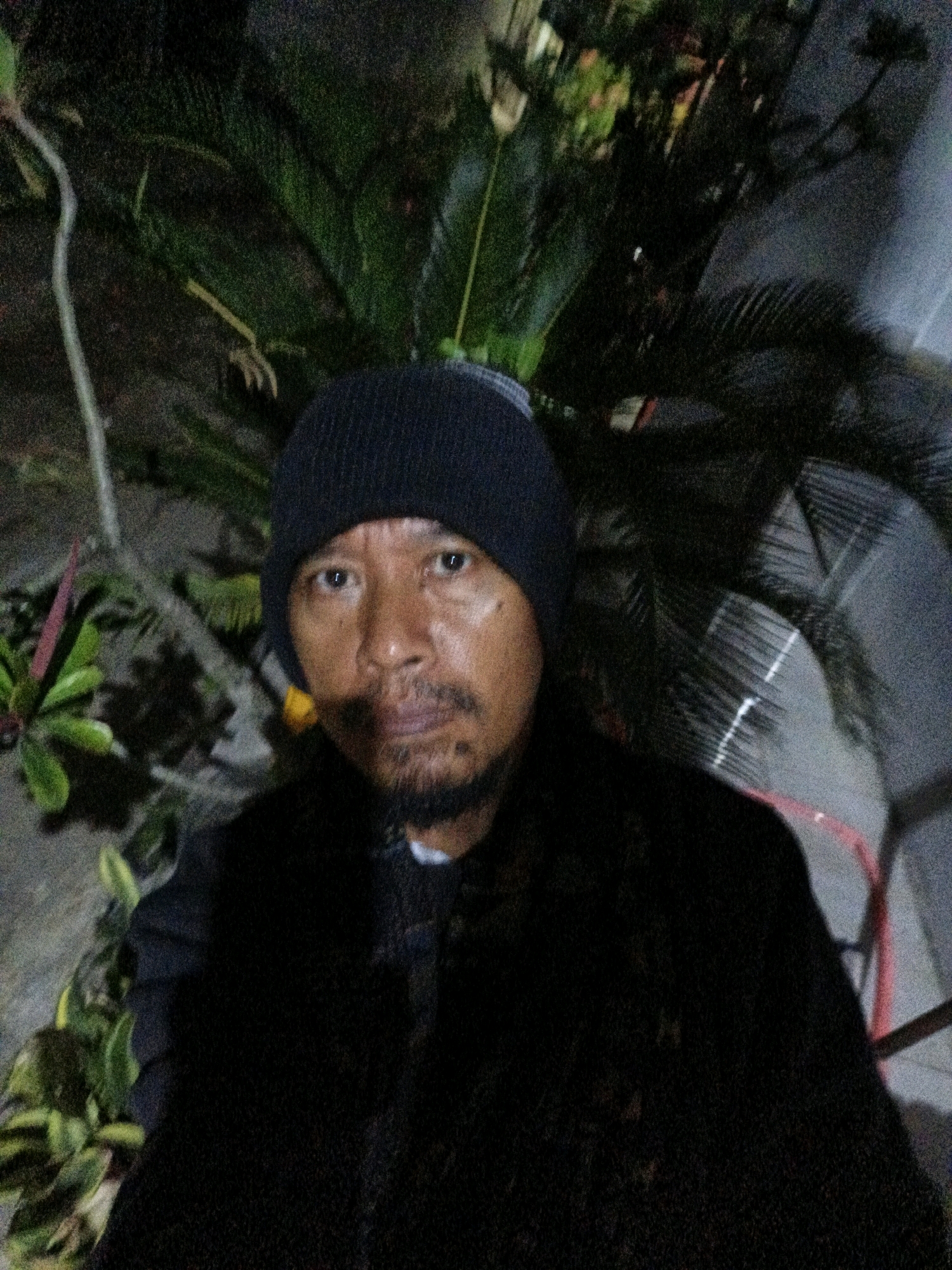 The image shows a man wearing a dark beanie and a dark jacket. He has a mustache and a beard. He is standing in front of some plants and what appears to be a wall or fence. The lighting suggests it is nighttime or dark. The man is the primary subject of the photograph, and he is looking towards the camera. There are no clear indications of the specific location within Tanahbaya, Indonesia, beyond the implied presence of vegetation and outdoor elements typical of such a region.