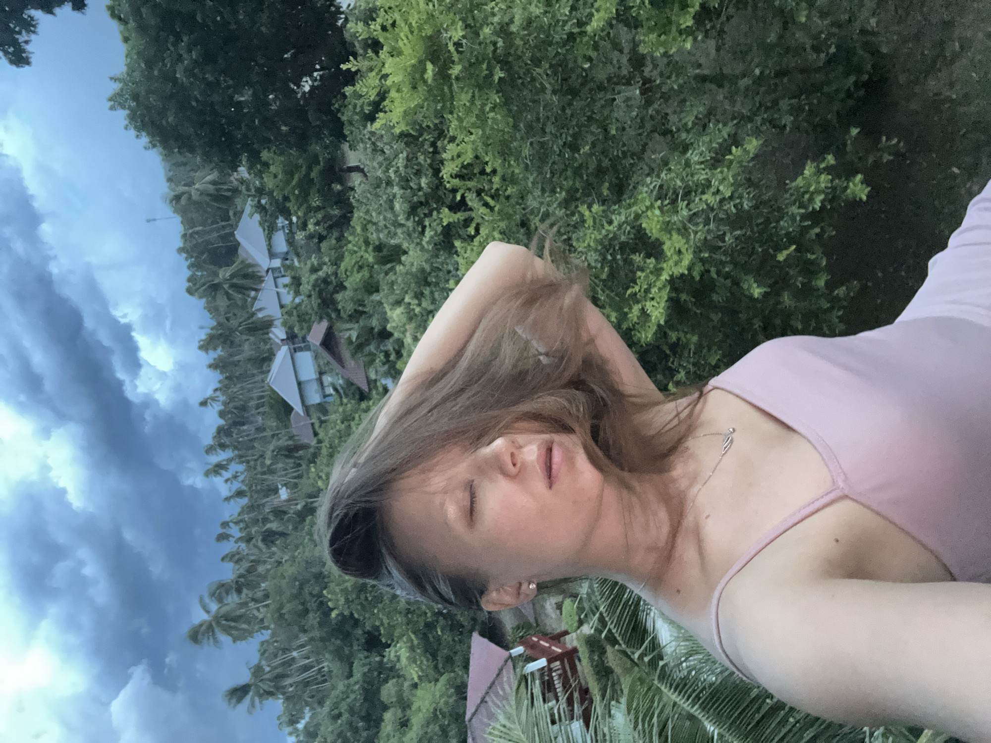 This is a self-portrait of a young woman with light brown hair, taken from a low angle. She is wearing a light pink tank top and a delicate necklace. Her eyes are closed, and her head is tilted back slightly, with one arm raised behind her head, suggesting a relaxed or perhaps contemplative pose. The background is a lush, green tropical landscape with palm trees and some buildings with red roofs, indicative of a resort or village setting. In the upper left portion of the image, a dramatic, cloudy sky hints at impending or recent rainfall, suggesting a late afternoon or early evening time. The overall atmosphere is serene and natural, possibly captured during a vacation or a moment of quiet reflection amidst a beautiful natural environment in Baan Hua Thanon, Thailand. There is no visible text in the image.