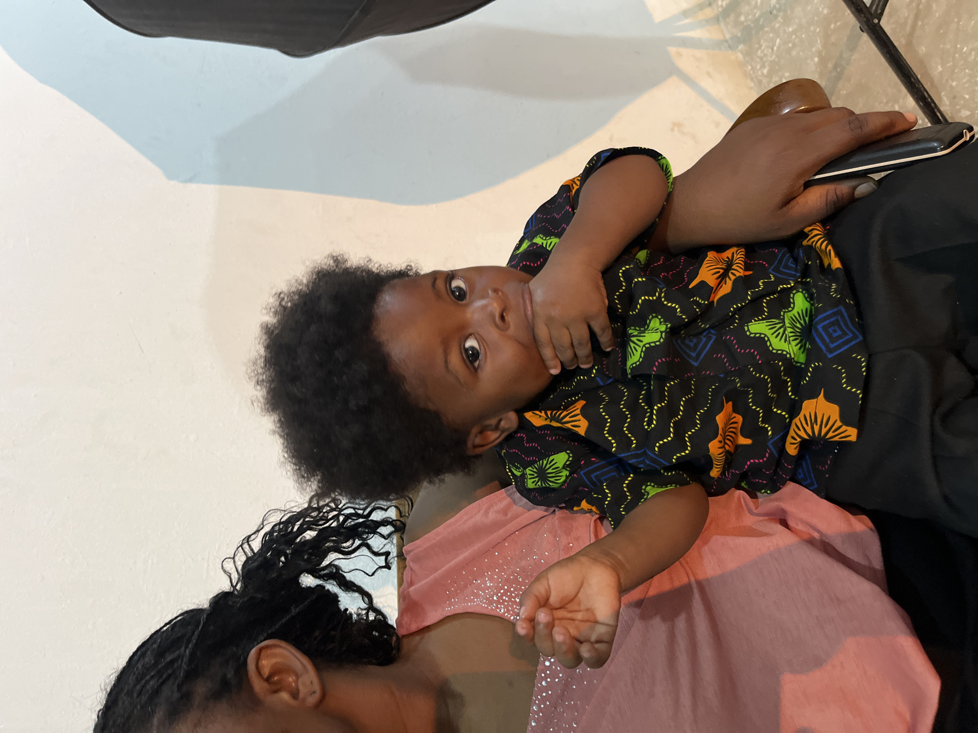 A young Black child with curly dark hair and wide brown eyes sits on the lap of a Black adult. The child wears a black shirt with a vibrant, abstract pattern of orange, green, and blue shapes. The child's right hand is near their mouth, and their left hand is resting on the adult's arm. The adult wears a shimmering pink top. The background is a plain, light-colored wall with a soft shadow on the right side. The scene appears to be indoors, likely in a residential setting in Gura Top, Nigeria.