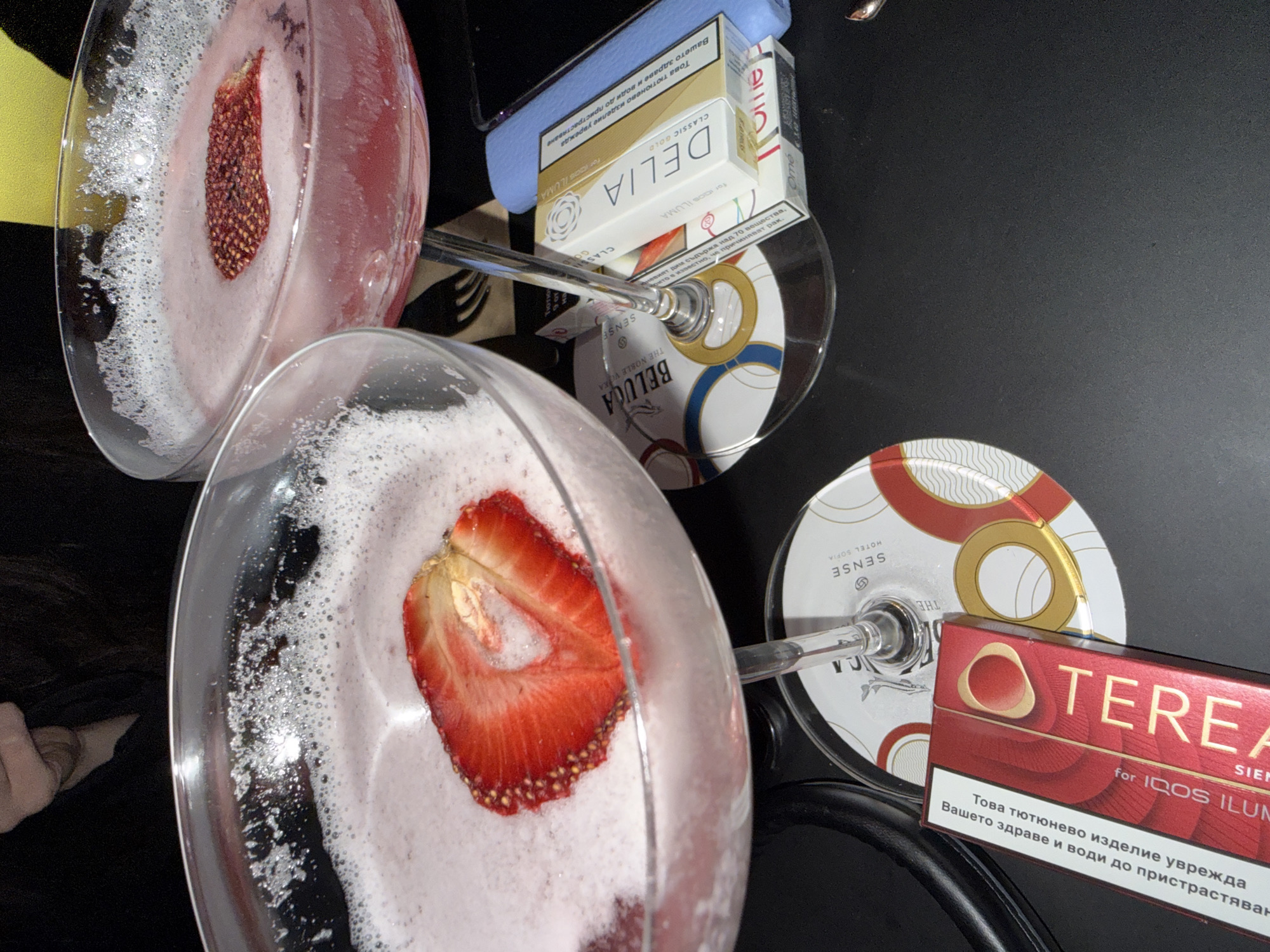 The image depicts two stemmed glasses filled with a frothy, pink liquid and strawberry slices. They are resting on a black surface amidst several objects. These include two boxes of "TEREA for IQOS ILUMA" and "DELIA CLASSIC GOLD for IQOS ILUMA" tobacco products, and two promotional coasters of a hotel named "SENSE HOTEL SOFIA". The tobacco products contain warning labels in the Bulgarian language. The setting seems to be indoors on a dark, flat surface.