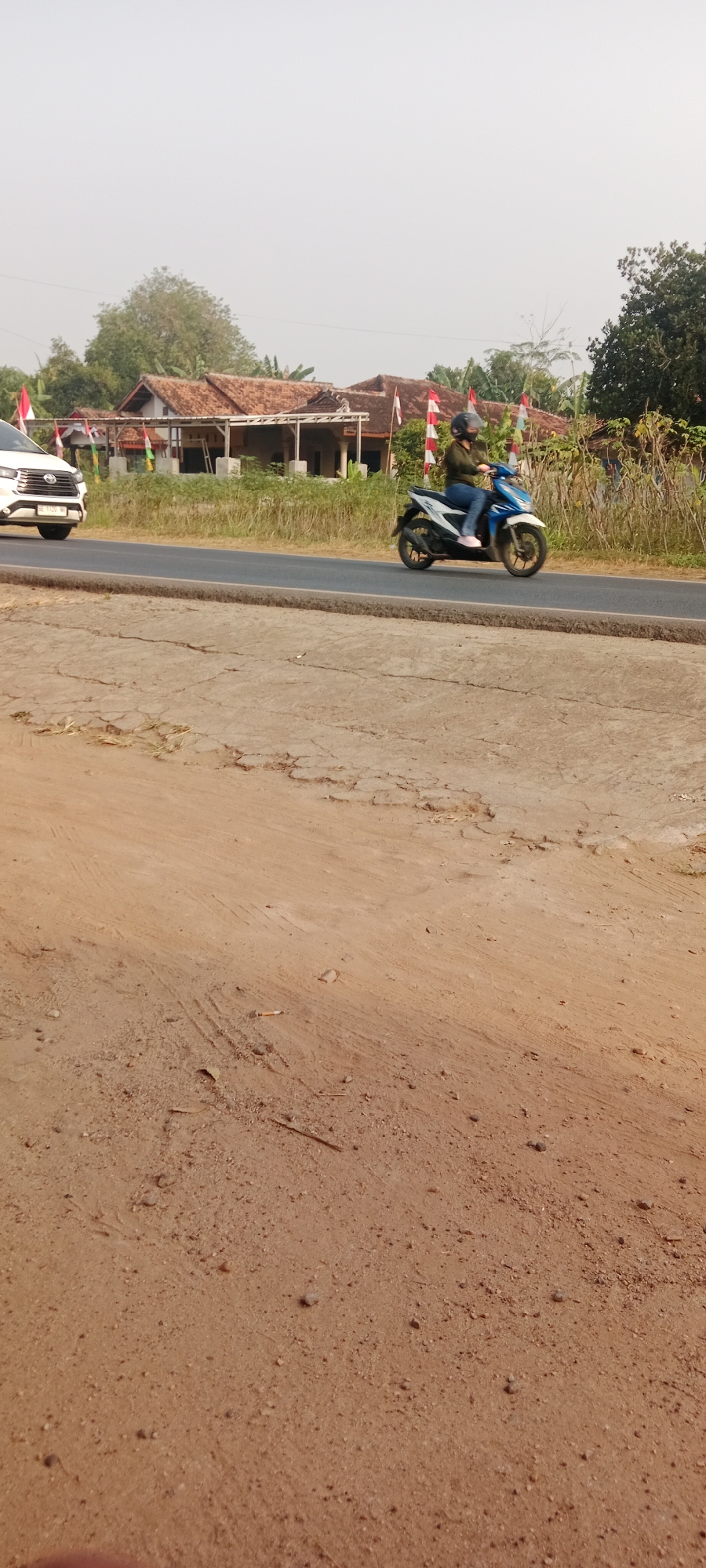 The image depicts a street scene in Gondang Rejo, Indonesia. In the foreground, a dry, cracked dirt surface with scattered small stones and debris is visible. A faint cigarette butt lies on the dirt in the lower center of the frame. Beyond this, a paved road runs horizontally across the image. On the road, a white SUV is partially visible on the left, and a person wearing a helmet and a dark green jacket is riding a blue and white motorcycle on the right, moving from left to right. The motorcycle rider is wearing blue jeans and pink shoes. In the background, a row of houses with red tile roofs and white pillars line the street. Red and white Indonesian flags are displayed on poles in front of the houses. Lush green vegetation, including trees and bushes, surrounds the houses and the road. The sky is a pale, hazy white, suggesting a bright but overcast day.