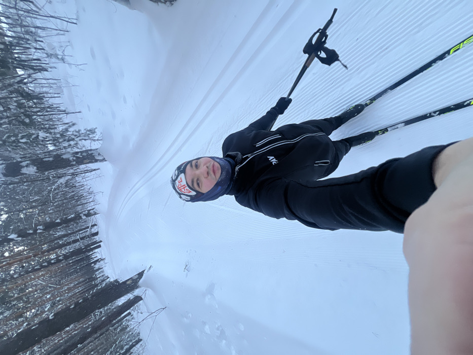 A lone individual is depicted in a wide-angle selfie while cross-country skiing on a groomed track within a snow-covered forest in Kazan, Russia. The subject, appearing male, is positioned centrally, looking directly at the camera. They wear a dark balaclava with red and white branding, a black athletic jacket featuring a half-zip, white vertical chest stripes, and "PAV" branding, along with dark gloves and pants. The individual is equipped with dark cross-country skis displaying yellow-green accents and partial "FISCHER" branding, and holds dark ski poles with beige grips. The track is covered in fresh snow, exhibiting distinct parallel grooves for classic skiing on the right side and a smoother section for skate skiing on the left. The environment consists of a dense forest with numerous tall, snow-laden conifer trees lining both sides of the ski path. The lighting is diffused, consistent with an overcast winter day. The camera is held by the subject's left hand, which is partially visible in the lower left portion of the image.
