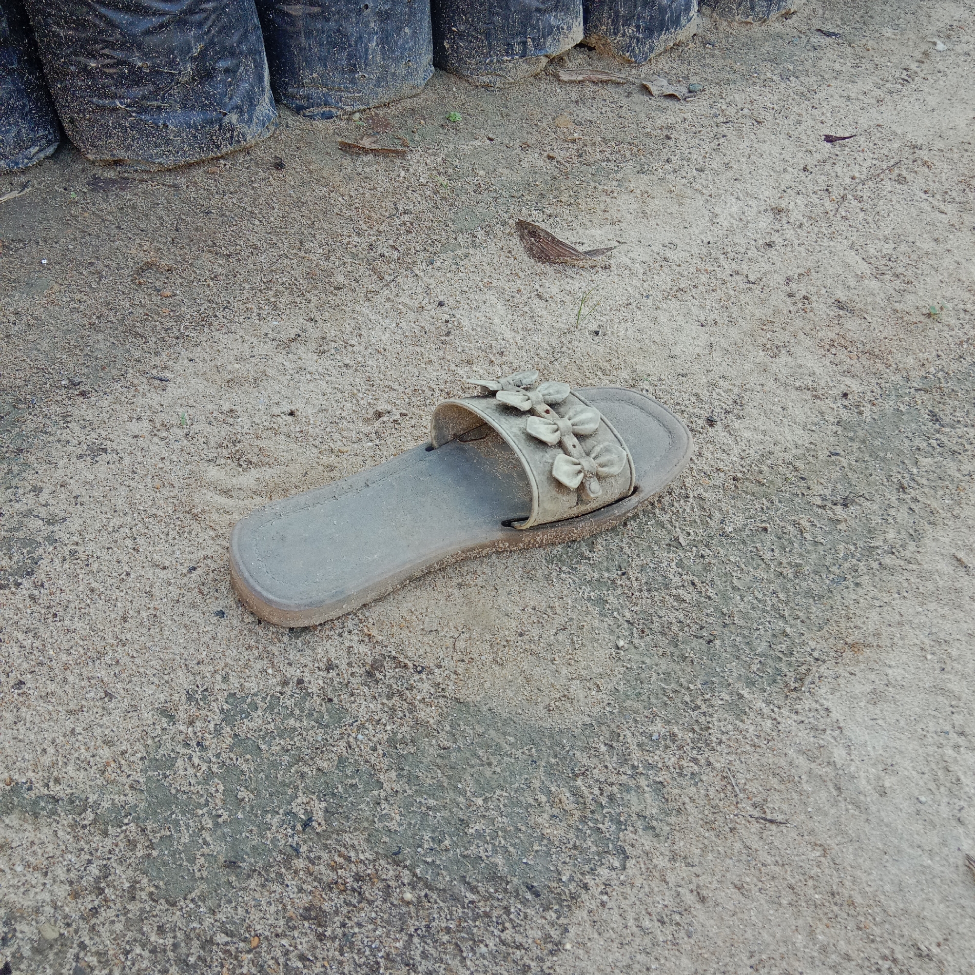A single, light-colored sandal lies on a sandy surface. The sandal has a decorative strap with three bow-like embellishments. In the background, several black plastic bags are lined up. The ground appears to be a mix of sand and some larger pebbles or debris, with a patch of darker, possibly damp earth visible. The scene is outdoors and appears to be in a dusty or unkempt area, possibly a garden or construction site. The image quality suggests it was taken with a mobile device. The specific location in Unknown, Indonesia is not discernible from the image alone.