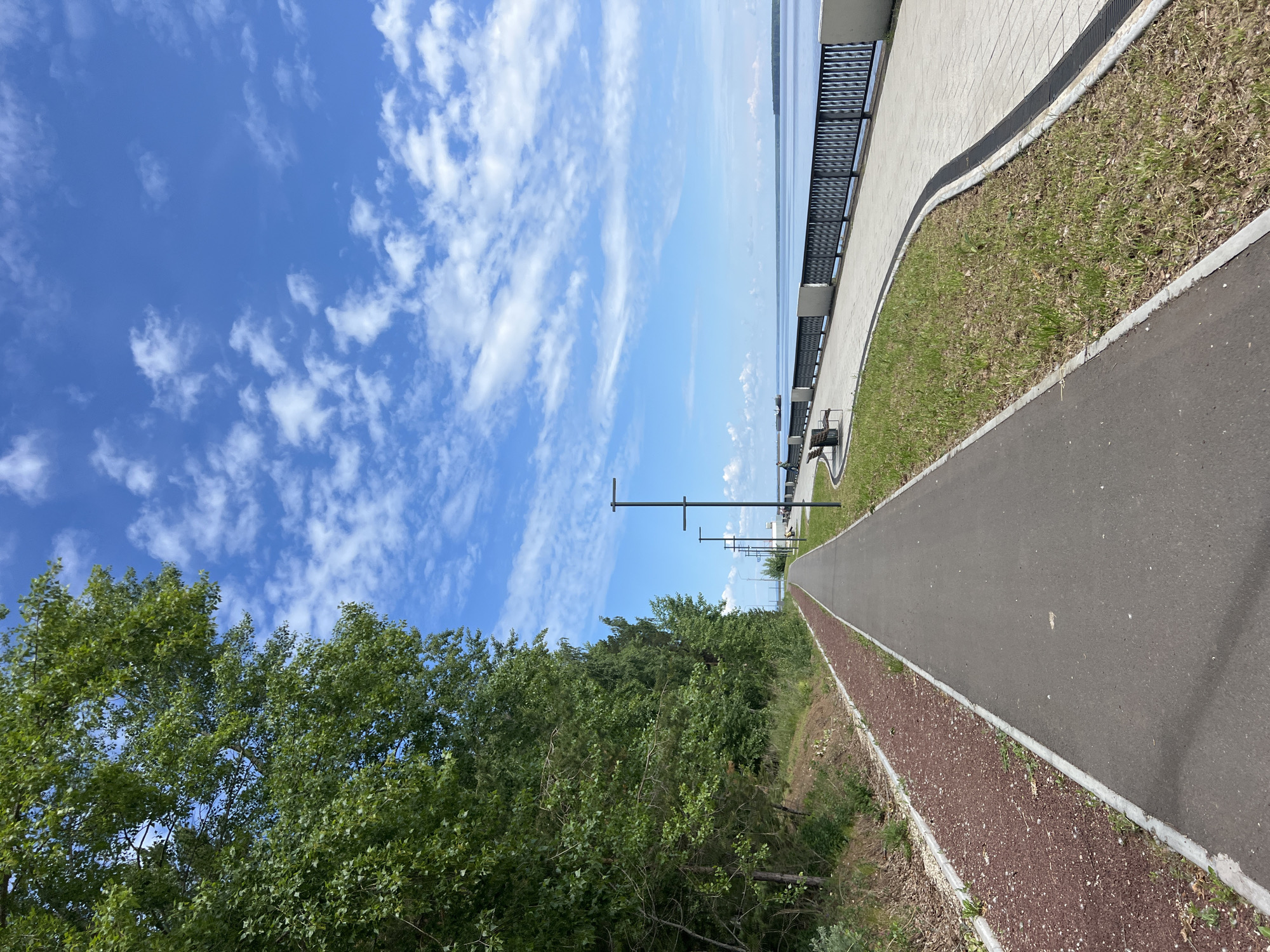 The image depicts a waterfront promenade in Chaikovsky, Russia, under a clear blue sky with scattered white cumulus clouds, indicating fair weather. The scene is bisected vertically by a long, straight path system.
On the left, a dense, verdant slope covered with deciduous trees and undergrowth rises steeply, forming a natural barrier. Adjacent to the base of this slope, a narrow, reddish-brown path with a textured surface runs parallel to the main thoroughfare, separated by a light gray curb. To its right, the primary path is a dark gray asphalt surface, also bordered by light gray curbs, extending into the distant perspective. One individual is visible walking on this asphalt path in the mid-ground, moving away from the viewer.
On the right side of the asphalt path, a narrow strip of grass with some sparse dry foliage separates it from a light-colored, paved promenade. A linear drainage grate runs along the edge of this paved area. The promenade is furnished with several dark-colored, modern benches facing the water. A line of identical dark-colored streetlights, featuring a single horizontal crossbar, extends along the promenade at regular intervals. A black metal railing with vertical bars and lighter-colored supporting pillars separates the promenade from a vast, calm body of water, likely the Kama River or Votkinsk Reservoir.
Two dark, low-profile vessels, resembling barges or cargo ships, are visible on the distant water surface. The far horizon reveals a faint, dark green line of land, indicating the opposite bank of the waterway. Further along the promenade, near the distant vessels, several figures are vaguely discernible.