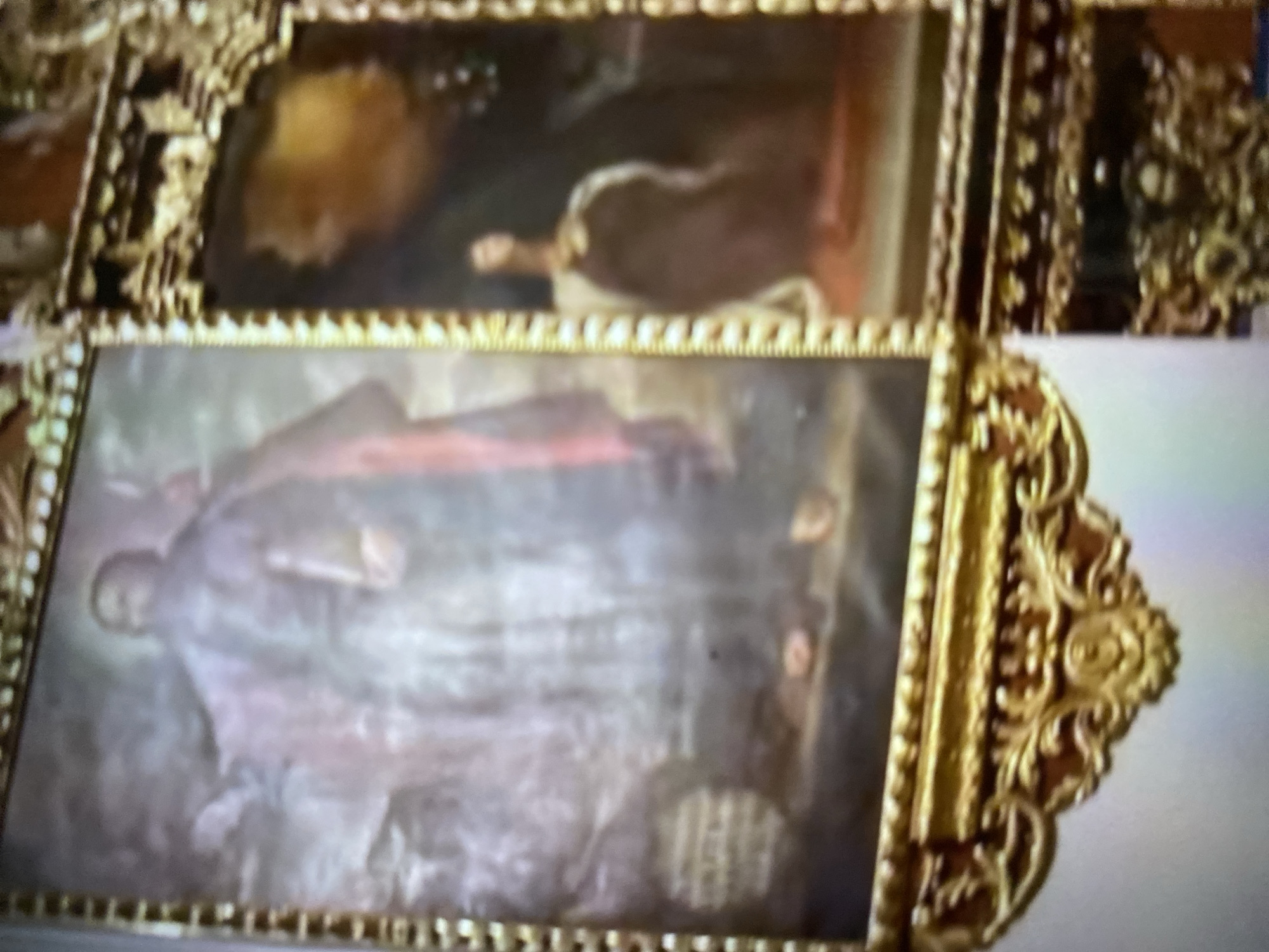 The image shows two framed paintings with ornate gold frames. The lower painting depicts a man in a flowing robe holding a book. The upper painting contains a figure in a dark robe. The location is likely the church in Tinaquillo, Venezuela.