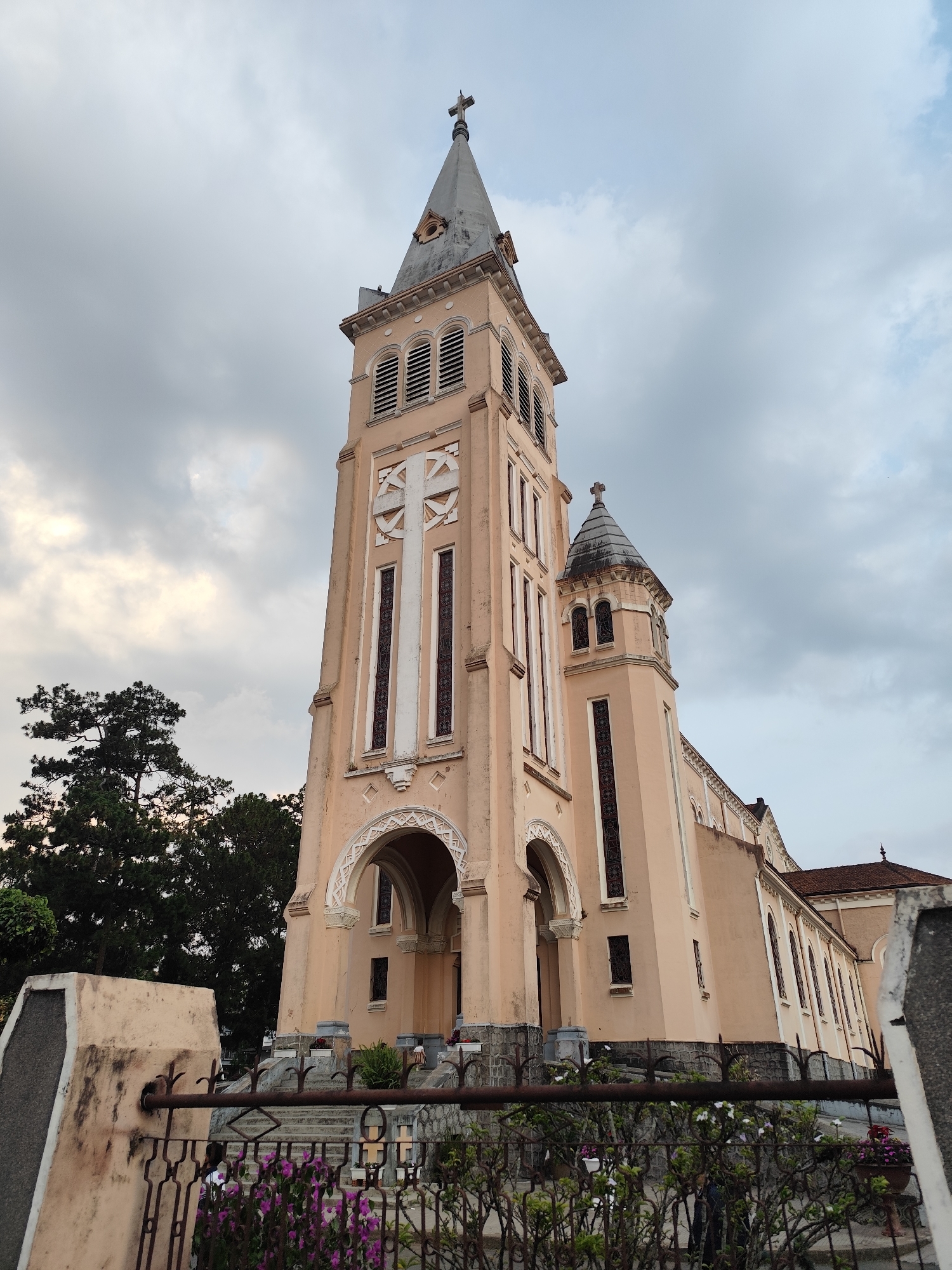 The image shows St. Nicholas Cathedral in Da Lat, Vietnam, taken from a low angle. The cathedral's peach-colored facade with white and brown accents is visible, along with its tall bell tower topped with a cross. The sky is overcast. A wrought iron fence and various plants are in the foreground. The tops of several trees are visible to the left of the cathedral.