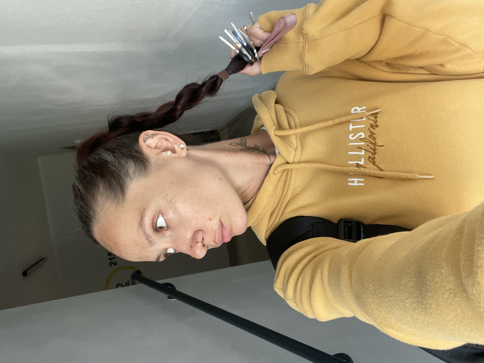The image shows a woman with a braid in her hair, holding a set of keys. She is wearing a yellow Hollister hoodie and a backpack. The woman has several piercings, including on her nose and ears, and a tattoo on her neck. She is looking to the side, with her mouth slightly open. The background appears to be a hallway with a light fixture and a sign that reads "215". The location is Pervomaysky, Russia.