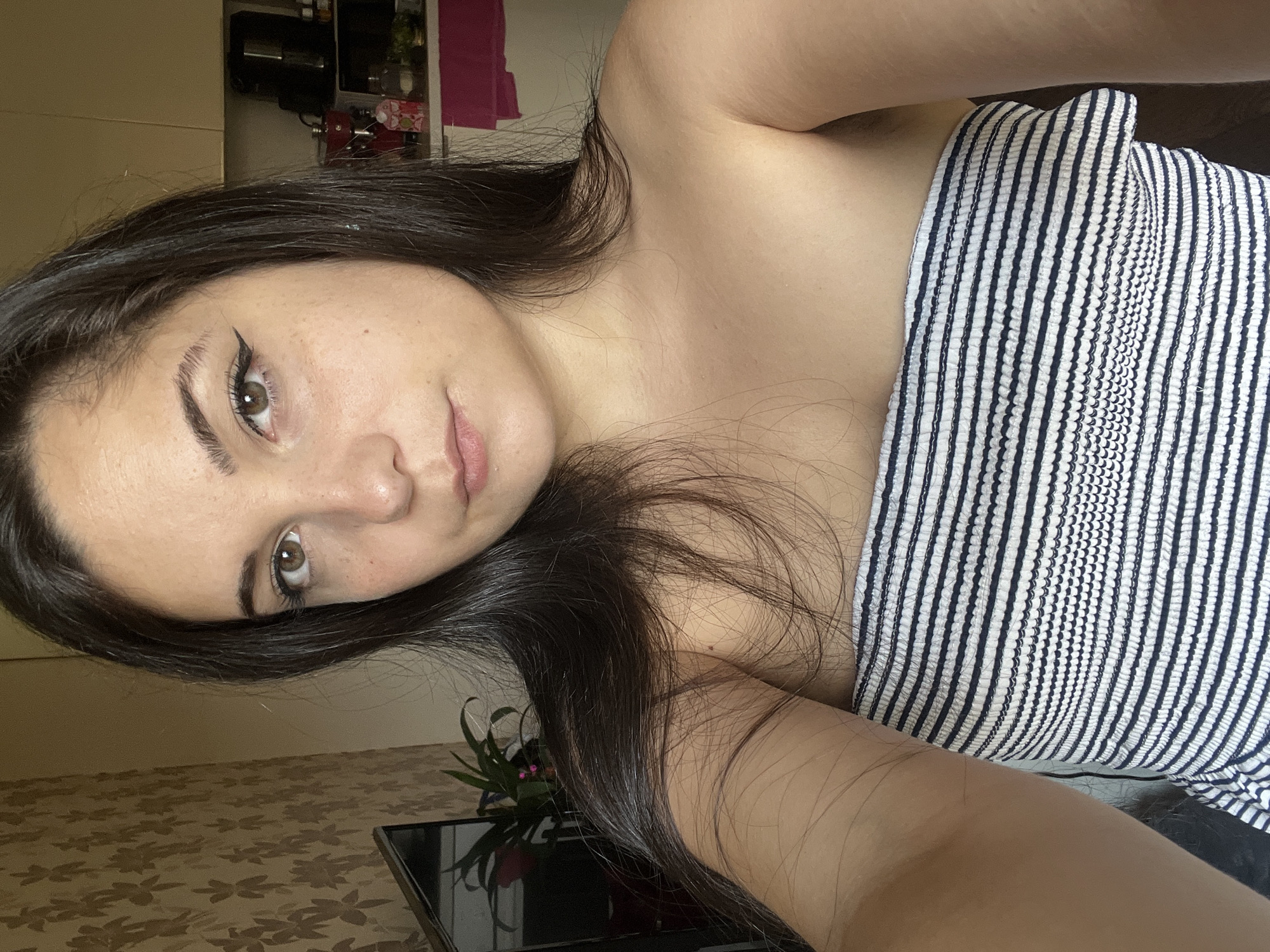 A young woman with long, dark hair and brown eyes is captured in a close-up selfie. She is wearing a strapless, horizontally striped top in navy and white. Her makeup includes winged eyeliner and mascara. The background appears to be an indoor setting, possibly a kitchen, with a tiled wall and some appliances visible. The lighting suggests natural daylight. The image does not contain any information about the city of Sofia, Bulgaria.