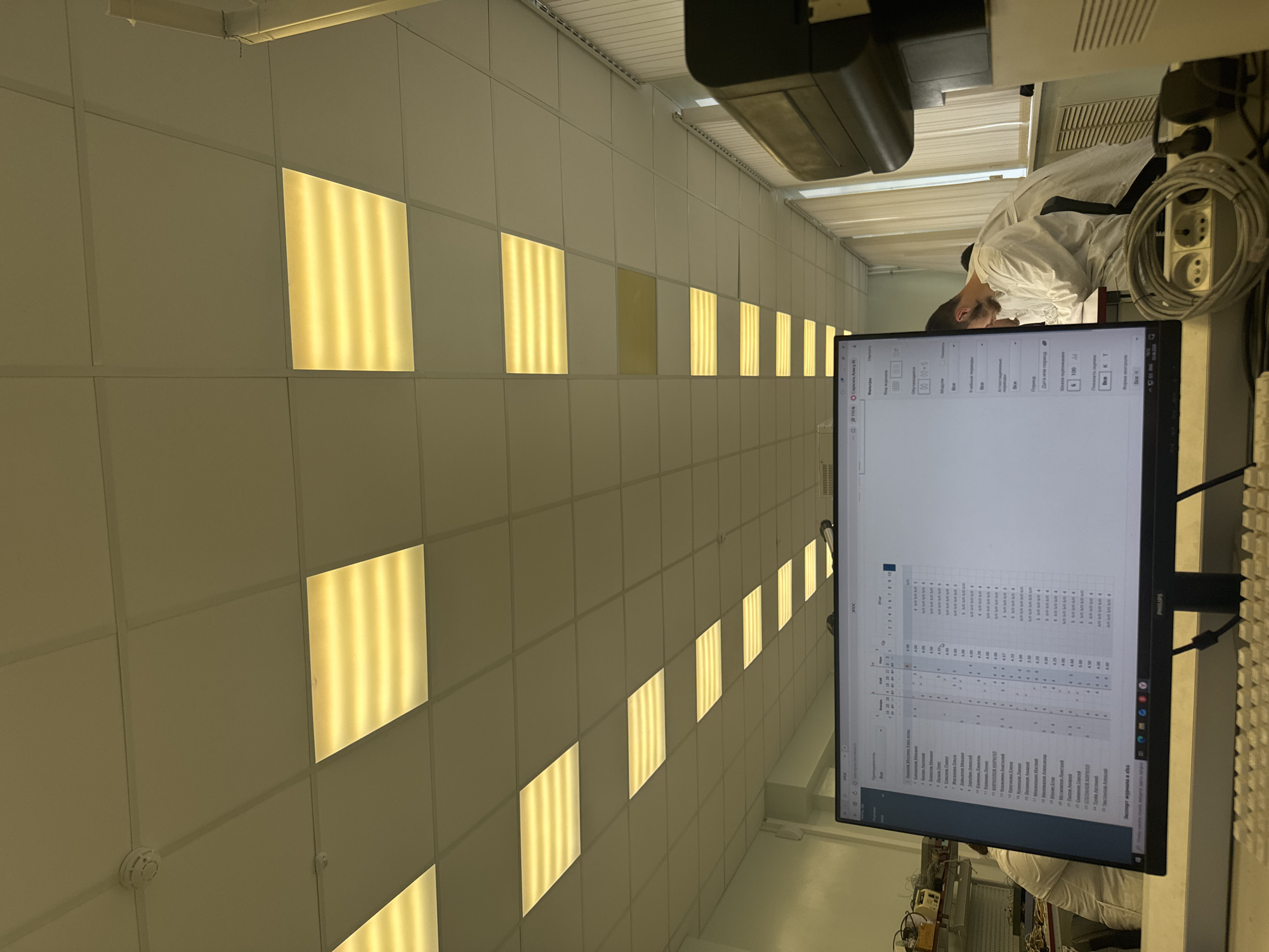 The image captures an indoor scene, likely a laboratory or cleanroom in Perm, Russia, characterized by white panel walls and a grid of uniformly spaced square light panels, some of which are illuminated with a warm yellow glow. A person, dressed in a white lab coat and sporting dark hair and a beard, is seated at a desk, intently looking at a Philips computer monitor.
The monitor displays a spreadsheet-like interface with multiple columns and rows of data. Visible Cyrillic text on the screen includes lists of names such as "Елисеев", "Жигалова", "Завьялов", "Зарубин", "Калинин", "КИРУЛЛОВ", "Лесников", "Механоши", "Милованя", "Симеонов", "СТЕПАНОВ", and "Маструбов". Other Cyrillic phrases visible are "Экспорт журнала в" (Export journal to), "Спросить Алику А" (Ask Alisa A, likely a virtual assistant prompt), "Фильтры" (Filters), "Период" (Period), "Дата или" (Date or), and "Все" (All). Numerical values like 10% and 100 are also present.
Behind the monitor, parts of a keyboard are visible. To the right of the person, coiled cables and power outlets are seen, along with what appears to be a printer or scanner on a shelf above. The room features a ventilation grate and a smoke detector on the ceiling. The environment appears clean and well-lit by artificial means, consistent with a professional or academic setting. The overall impression is one of focused work or study.