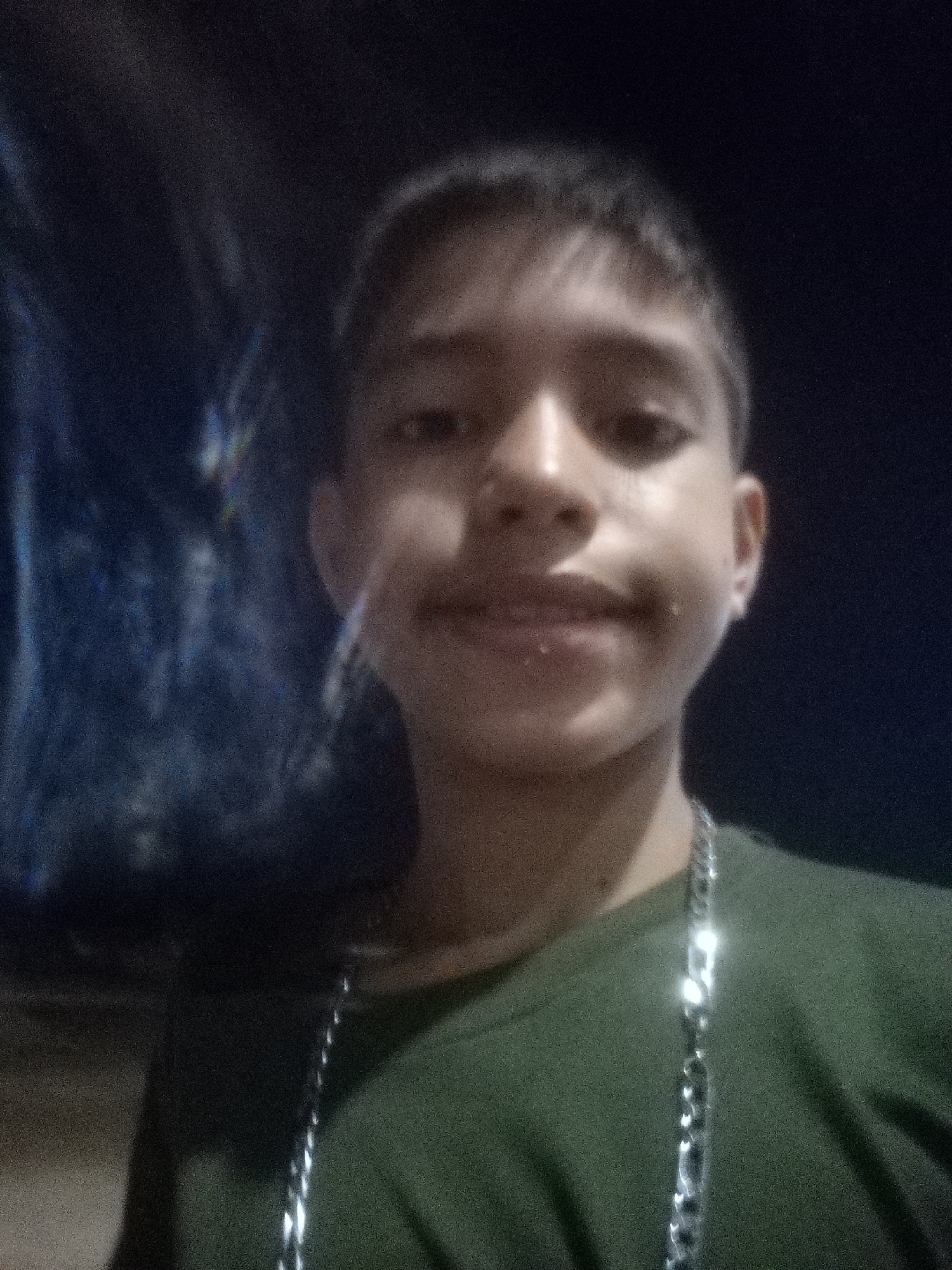 A close-up, dimly lit selfie of a young person with short dark hair and a dark green t-shirt. They are wearing a silver chain around their neck. The background is very dark, possibly nighttime, with indistinct shapes that may suggest a street scene. No specific details confirm the location as Juan Griego, Venezuela.