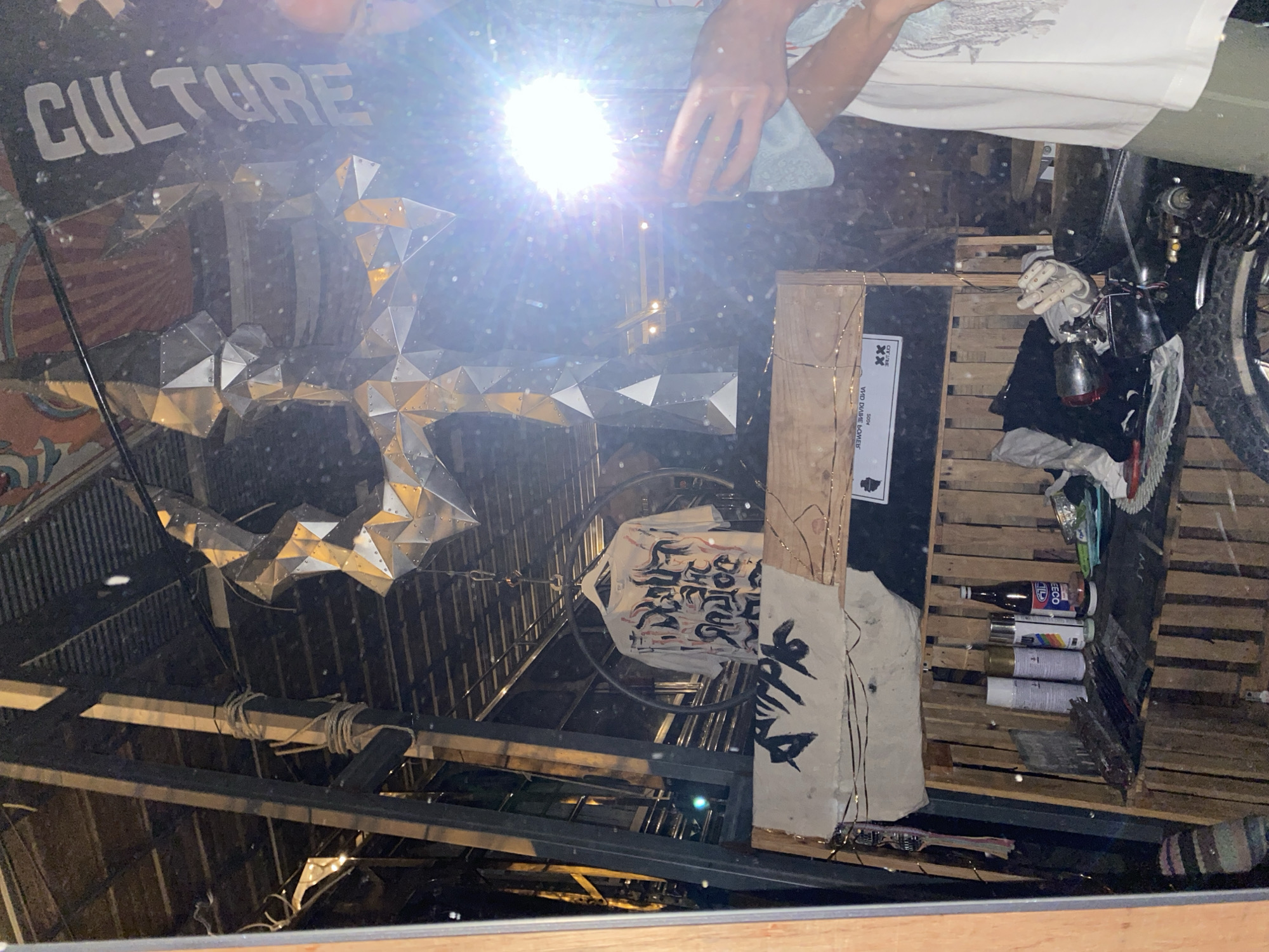 The image shows a cluttered, reflective interior space with visible ceiling beams and an irregular, metallic sculpture. A sign reading "CULTURE" is partially visible. A person holding a reflective object is present, creating a bright glare in the center. The space contains a motorcycle, spray paint cans, clothing, and wooden pallets. The location is Baan Nai Suan, Thailand.