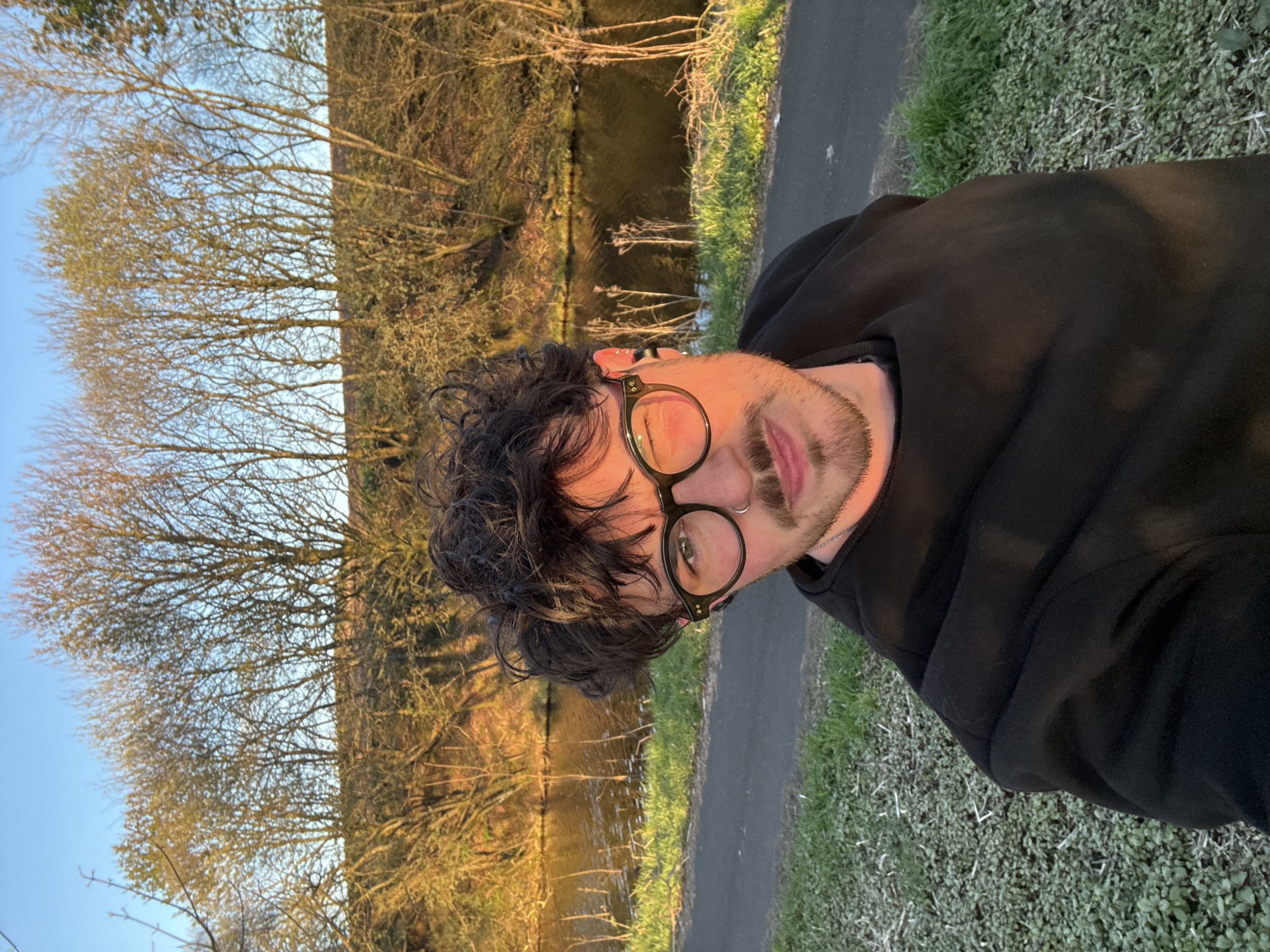 A young man with dark, slightly messy hair and a short beard looks directly at the camera in what appears to be a selfie. He wears dark-rimmed, round glasses and a subtle nose ring (septum piercing). His expression is neutral or slightly pensive. He is dressed in a plain black hoodie or sweatshirt, with a thin silver chain visible around his neck.
The setting is outdoors during what appears to be the golden hour of late afternoon or early evening, evidenced by the warm, directional sunlight. Behind the man, a calm body of water, likely a canal or narrow river, stretches horizontally across the frame. On the far bank of the water, a dense line of bare deciduous trees with intricate branches catches the warm light. The presence of bare branches suggests late autumn, winter, or very early spring.
To the man's right, a narrow paved path runs alongside a strip of green grass and low-growing foliage. The sky above the trees is clear and blue. The warm sunlight casts a glow on the man's face and the landscape, creating reflections on the water's surface. There is no visible text in the image.