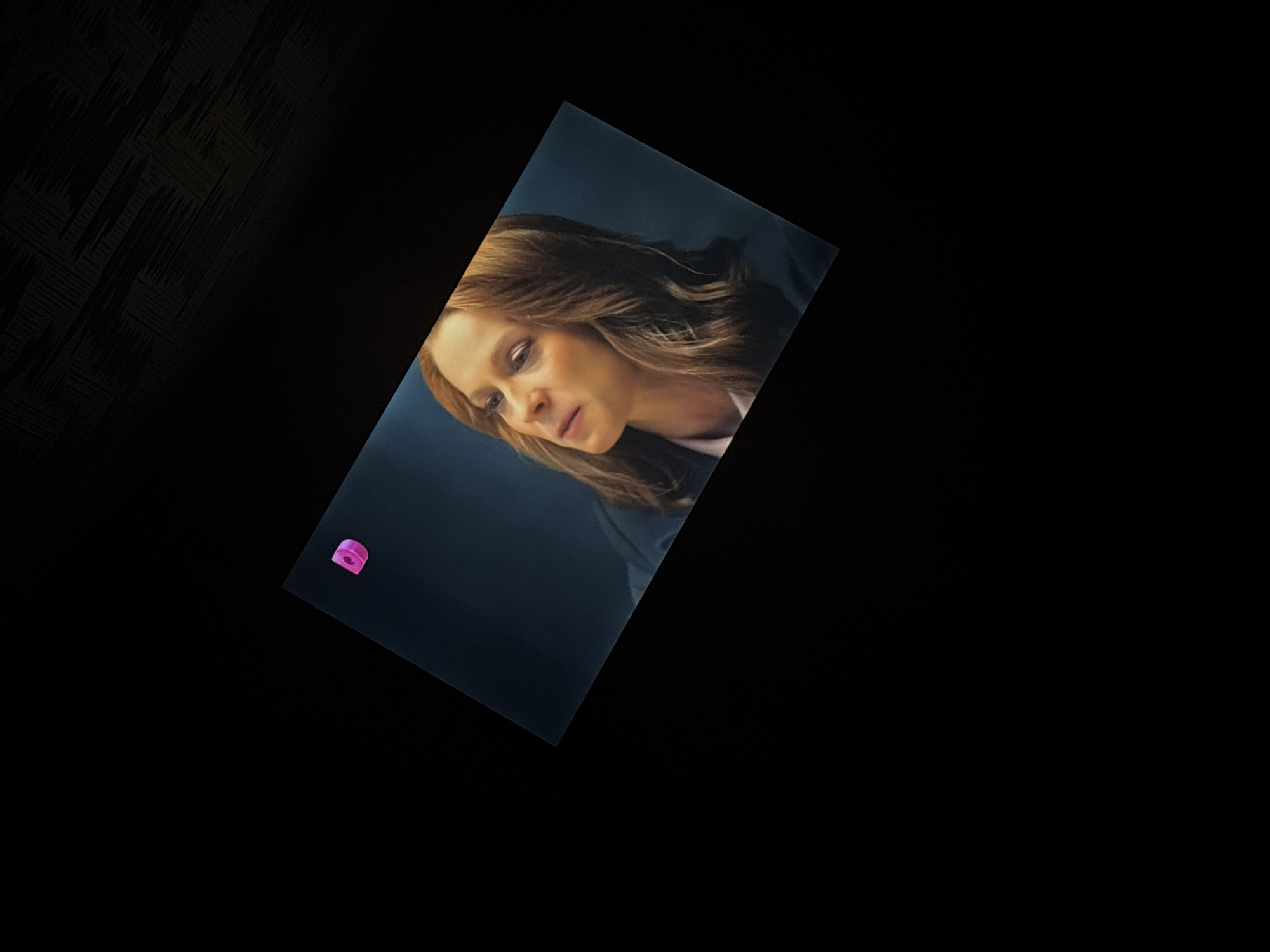 The image shows a close-up, angled shot of a television screen displaying a scene from a show. The screen is black with a pink logo in the upper left corner. The content on the screen features a woman with auburn hair and makeup, wearing a dark jacket over a white shirt. She is looking to her left with a thoughtful expression. The background of the screen is a dark blue. The television screen is tilted and appears to be viewed in a dark room, with only the screen being illuminated. The dark background of the room has a textured pattern visible in the upper right corner.