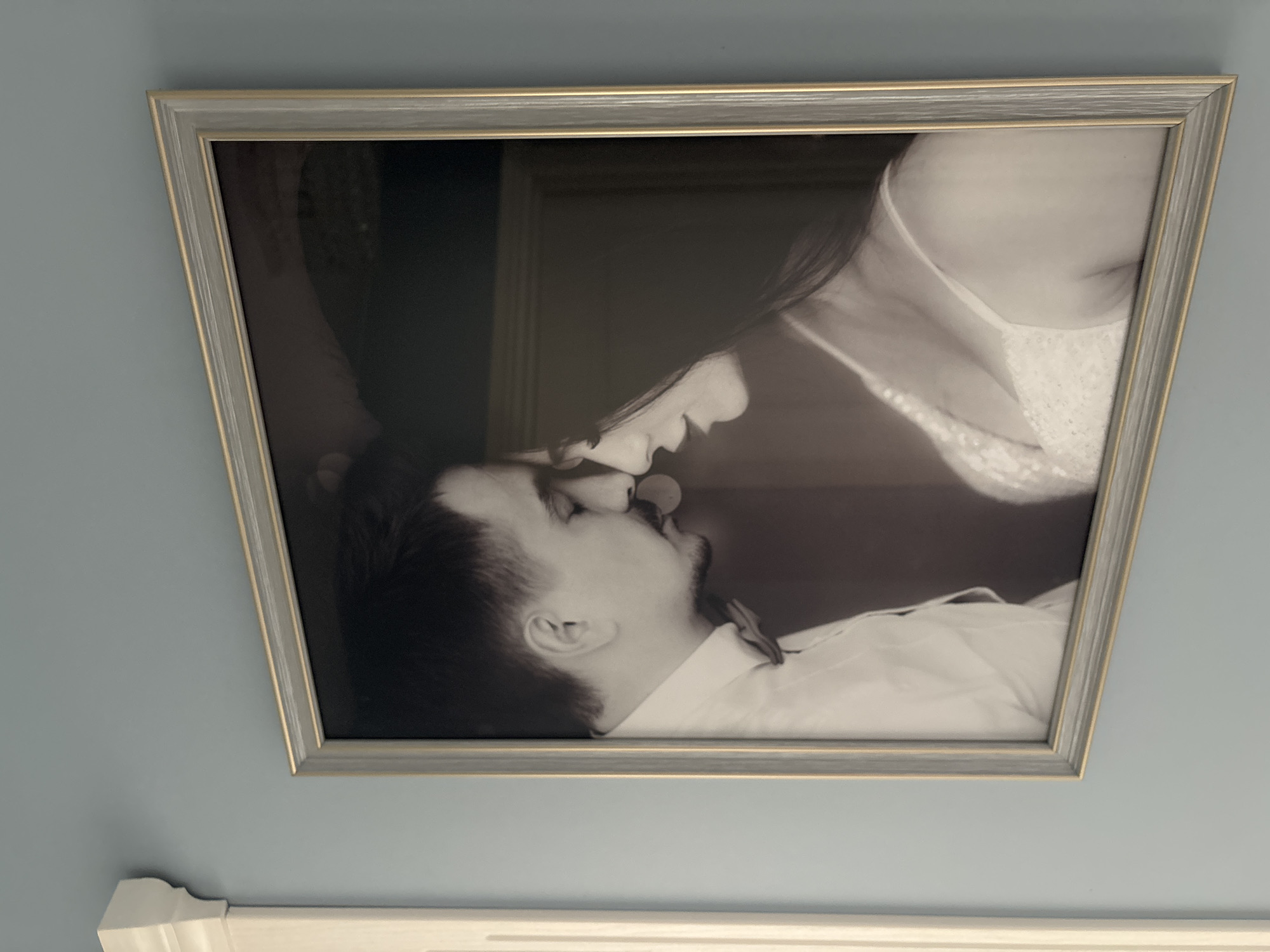 A framed black and white photograph hangs on a light blue wall above a white decorative molding. The photograph depicts a couple embracing and nearly kissing. The person on the left appears to be male, with short dark hair and a trimmed beard, wearing a light-colored dress shirt. The person on the right seems to be female, with long dark hair, wearing a light-colored garment with lace detail. The frame is silver-grey with a thin gold accent on the inner edge. The location is indoors and the city/state are unknown.