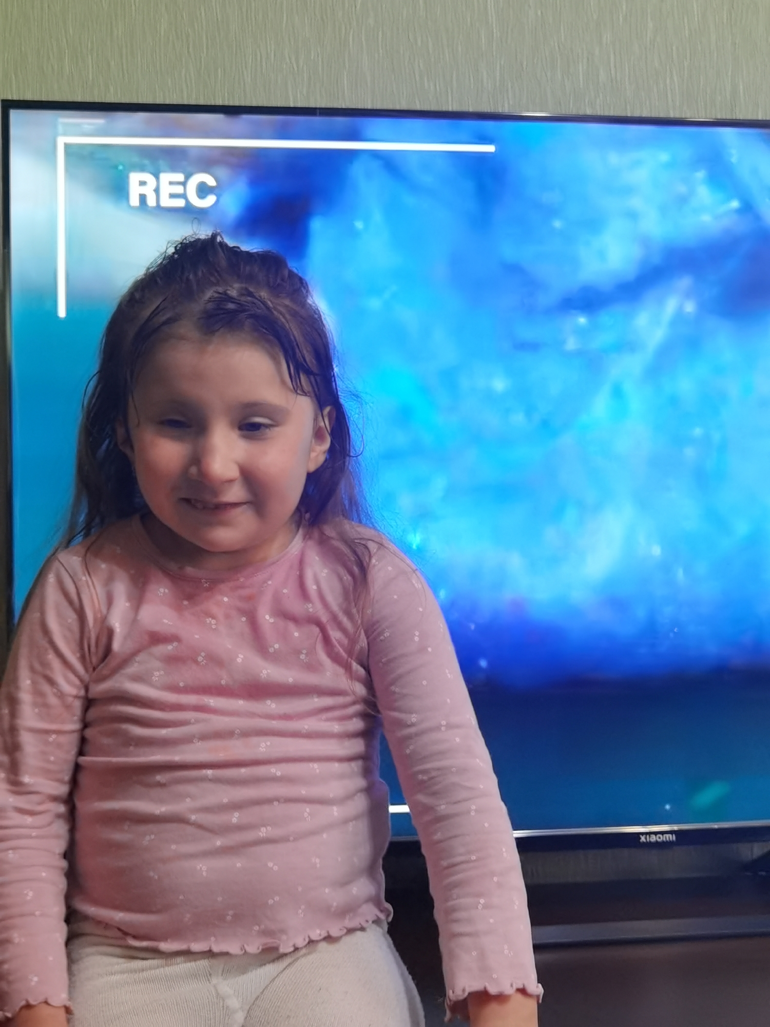 The image depicts an indoor scene, described as being in the city of Chaikovsky, Russia. A young girl, appearing to be Caucasian, is centrally positioned, looking directly at the viewer with a slight smile. She has dark, unkempt hair that is parted and falls past her shoulders. She wears a light pink, long-sleeved top with a ruffled hem, adorned with small white floral or polka dot patterns. Light-colored (possibly off-white) leggings or thermal pants are visible below her waist. Behind the girl, a large flat-screen television is active, displaying an abstract, vibrant blue and teal image. In the upper left quadrant of the TV screen, a white on-screen display (OSD) overlay shows a rectangular frame with a vertical line extending downwards, enclosing the white text "REC." The brand "XIAOMI" is visible in white lettering on the lower right bezel of the television. The wall behind the TV and the girl features a light-colored, vertically textured wallpaper. A dark, flat surface, likely a television stand or table, is situated beneath the TV.