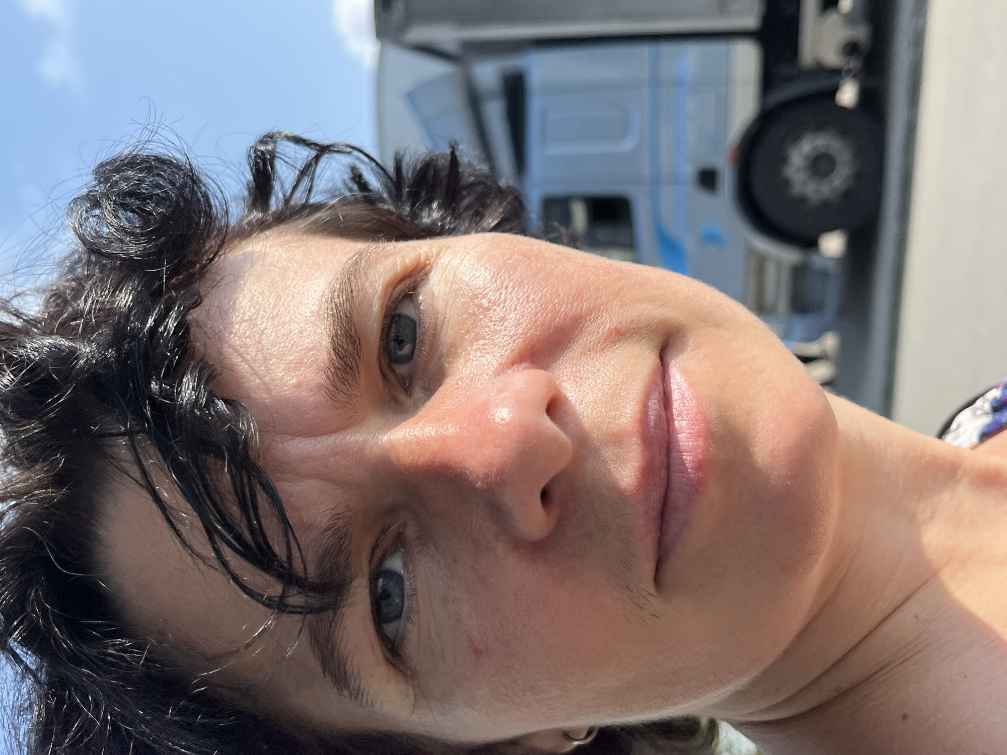The image shows a close-up, outdoor, waist-up portrait of a person with dark, curly hair and fair skin. The person's face is illuminated by sunlight, with shadows cast on the left side of their face and neck. The person is looking directly at the camera with a neutral expression. Their eyes are blue, and they have a small mole on their left cheek. They are wearing a silver hoop earring in their left ear. In the background, out of focus, is a large, light-colored truck, suggesting an urban or industrial setting, possibly a roadside location. The sky is blue with some clouds. The image appears to be taken on a sunny day. The location is specified as Ureki, Georgia.