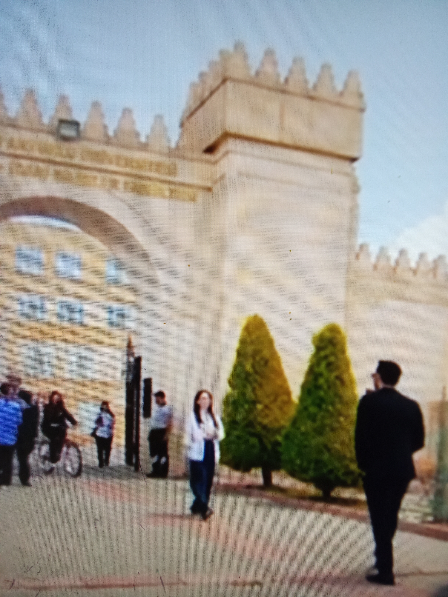 A group of people are seen in front of an arched entrance to a building. The building appears to be made of light-colored stone and has crenellated battlements along the top. Text is visible above the arch, partially obscured by the battlements. Through the arch, windows of a building can be seen. Two conical trees stand in front of the building, to the right of the arch. On the left side of the image, a person is riding a bicycle. Other individuals are standing or walking near the entrance. The ground is paved with cobblestones. The sky is a pale blue. The scene appears to be a public space, possibly a university or government building. The location is identified as Рощин ауылдық округі, Kazakhstan.