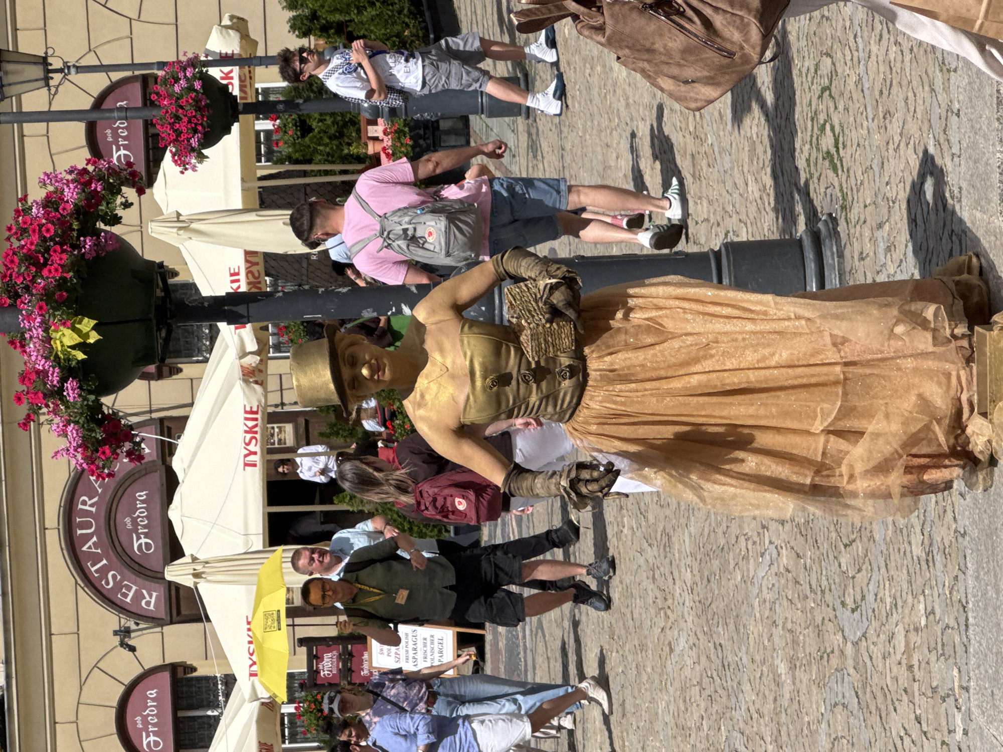 The image depicts a bustling outdoor scene in front of "RESTAURACJA pod Fredra" in the Market Square (Rynek) of Wrocław, Poland. The focal point is a person performing as a living statue, entirely painted in gold, positioned slightly right of the frame's center. This individual wears a gold top hat, a gold corseted bodice with three rose-like embellishments on the right side, long gold gloves with ruffled cuffs, and a sheer, flowing gold skirt. They hold a small, textured, gold-colored basket in their gloved hands.
In the background, numerous pedestrians walk on a cobblestone surface. Visible individuals include:
* A man in a dark green short-sleeved shirt and dark shorts, holding a yellow umbrella, walking towards the viewer's left.
* A man in a pink t-shirt and blue denim shorts, wearing a grey backpack, walking away from the viewer.
* A man in a white t-shirt, grey shorts, and sunglasses, walking towards the viewer's right.
* A woman in a blue shirt and white shorts near the left frame edge.
* A woman in a patterned top and jeans carrying a red backpack.
The restaurant building, constructed of light-colored stone, features an arched entrance with "RESTAURACJA" and "pod Fredra" signs in red lettering. Several large, cream-colored outdoor umbrellas with red "TYSKIE" logos are set up in front of the restaurant, providing shade for outdoor seating. Two dark lampposts, one near the golden statue and another further back, are adorned with large hanging baskets overflowing with vibrant pink and red flowers. A white signboard, partially obscured, displays "ŚWI POL ASPARAGUS" and "FRISCHER POLNISCHER PARGEL." The scene is illuminated by bright sunlight, casting distinct shadows on the cobblestone ground.