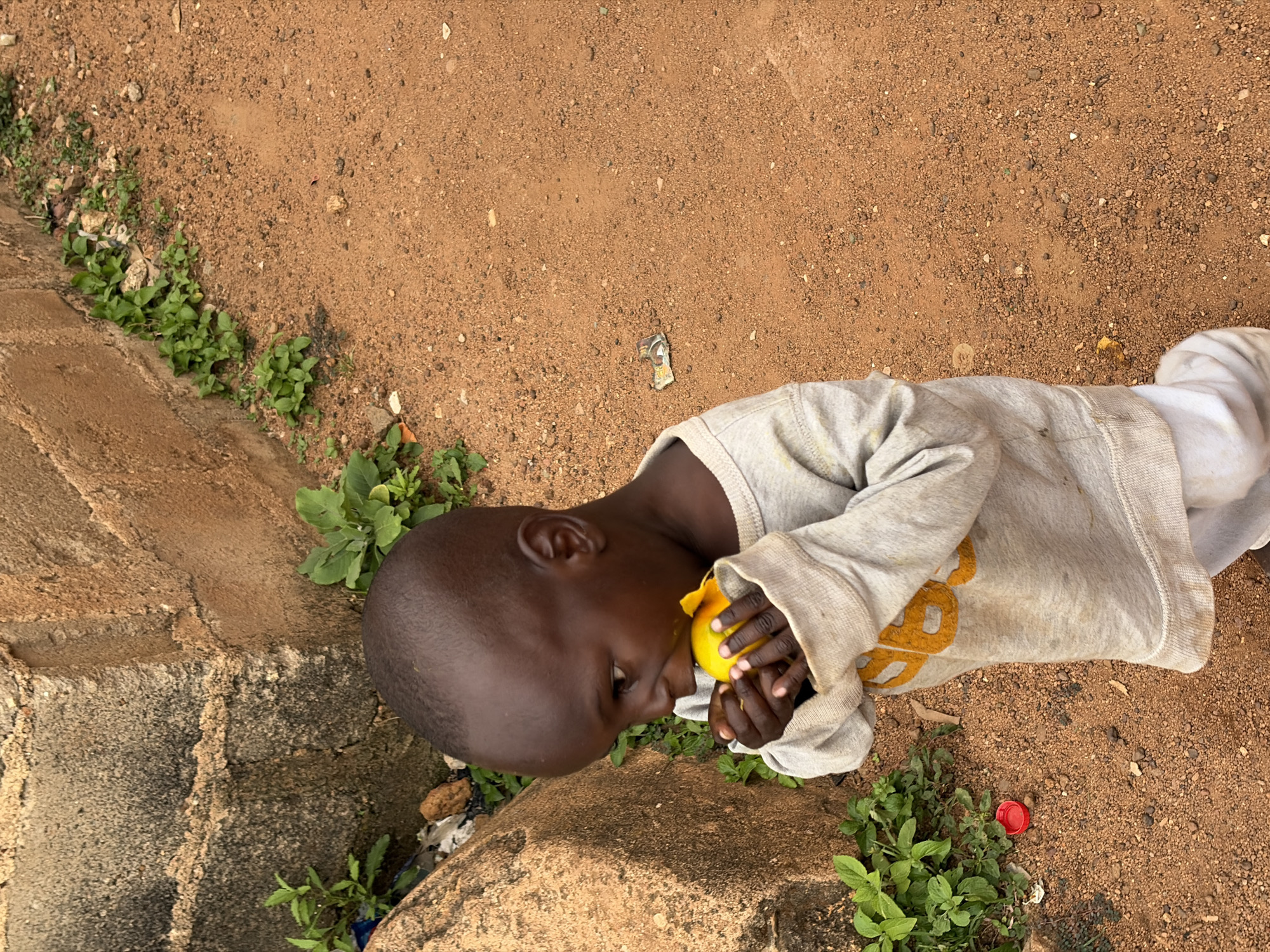 A child is lying on the ground in an outdoor area. The ground is mostly reddish-brown dirt, with some green vegetation growing near the edge of a concrete or stone structure. The child is dark-skinned and wearing a gray long-sleeved shirt and white pants. The child is holding a yellow fruit or object near his face. In the background, the ground is bare dirt with small pebbles and some litter, including a red bottle cap. The child is in the city of Barakin Lerawi, Nigeria.