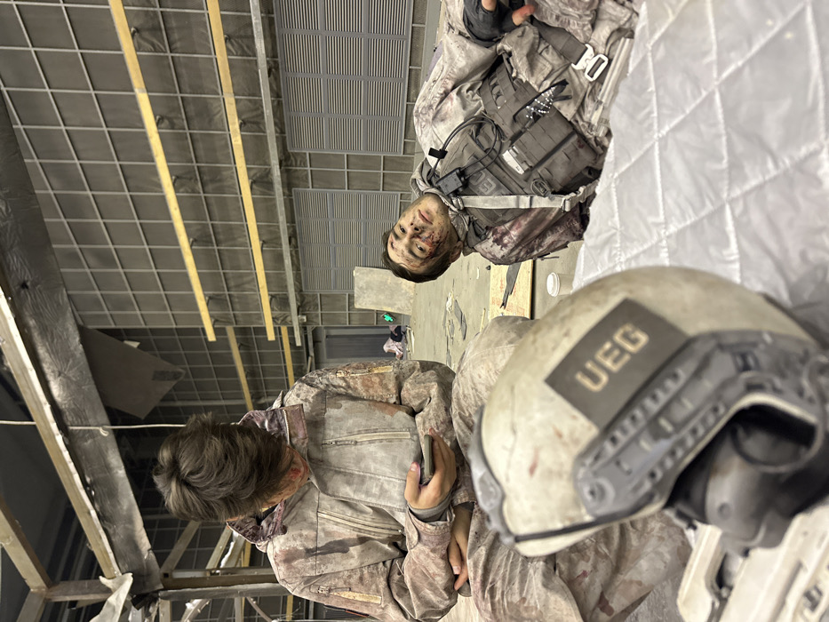 The scene depicts an indoor environment, likely a film set or large studio, situated in Huangdao District, China. The background features a grid-patterned wall system with horizontal support beams and structural elements, with scattered debris on the floor. Two male individuals, both exhibiting simulated bloodstains on their clothing and faces, are present.
On the left, a male subject with dark hair is seated on the floor, wearing a light-colored camouflage-patterned tactical suit. His face and suit are marked with simulated blood. He is looking down and actively interacting with a smartphone held in both hands.
On the right, a second male subject, also with dark hair, lies reclined on a quilted light-colored mat. He wears a similar blood-stained camouflage uniform underneath a dark tactical vest equipped with multiple pouches and a communications device attached to his shoulder strap. Simulated blood is visible on his face, particularly around the eyes and cheek. He has open eyes and a neutral expression, looking towards the viewer.
In the foreground, a light-colored tactical helmet, also bearing simulated bloodstains, is visible. The helmet features a dark rectangular patch on its side inscribed with the letters "UEG". A small white cup rests on the floor near the reclining individual.