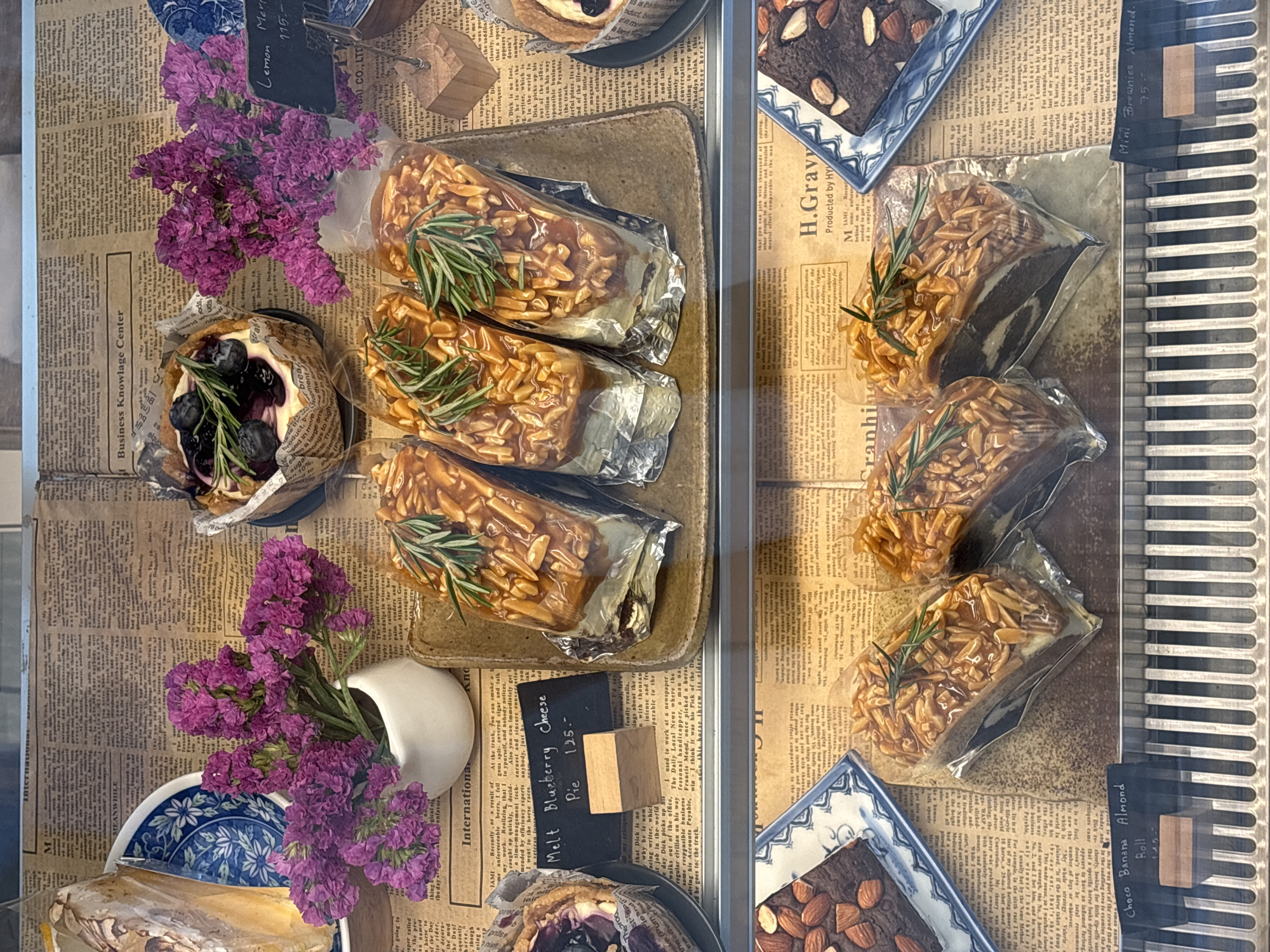 A display case in a shop in Choeng Thale, Thailand, features an assortment of pastries. On the left are blueberry cheese pies and almond pastries, beside purple flowers in a white vase on a blue and white plate. On the right are almond and brownie pastries, each individually wrapped. Small black signs with white writing and wooden stands identify the pastries and their prices. The display case's bottom right edge has a metal grill. The backdrop is newspaper print.