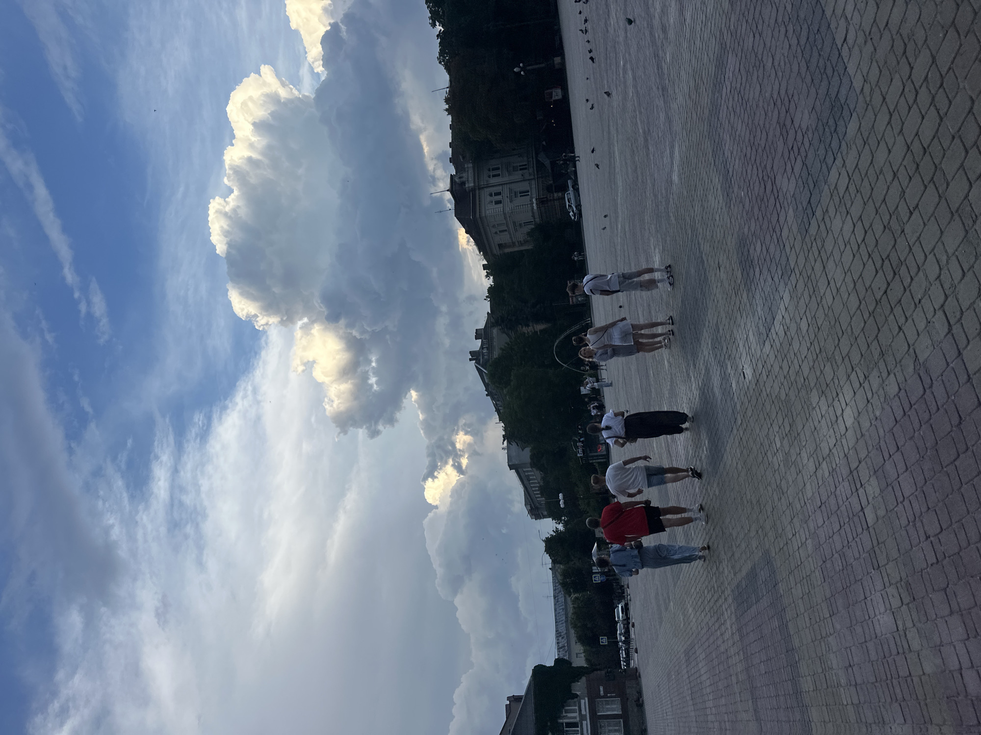 In Ternopil, Ukraine, a group of five individuals walks across a wide, paved plaza. The sky above is dramatic, with a mix of dark, heavy clouds and bright, puffy white clouds illuminated by the sun, suggesting either early morning or late afternoon. The ground of the plaza is made of interlocking gray and red pavers laid in a uniform pattern.
The group consists of three men and two women. From left to right, the first person is a woman in a denim jacket and light-colored pants. Next to her is a man wearing a red t-shirt and dark shorts. Following them is another man in a white t-shirt and light-colored shorts. The fourth person is a woman in a white top and patterned skirt, and the last person in the group is a man in a white t-shirt and light-colored shorts, carrying a bag. They are walking away from the camera.
In the background, buildings of various architectural styles line the edge of the plaza, some appearing to be older, while others are more modern. Trees are visible between and behind some of the buildings, adding a touch of greenery to the urban landscape. A few cars are parked on a street beyond the plaza. Pigeons are scattered across the paved area, some foraging. The overall scene captures a moment of people moving through an open urban space under a dynamic sky.