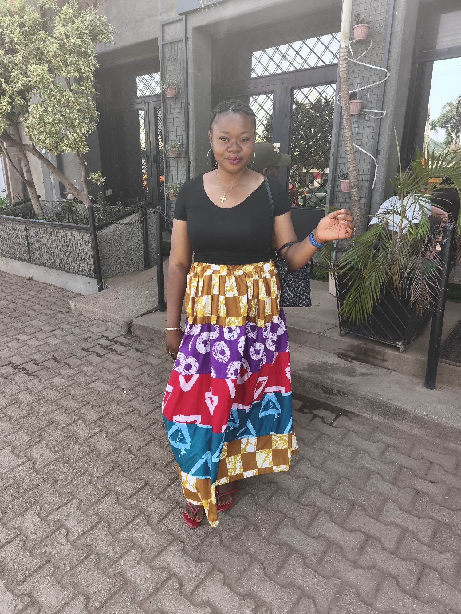 An outdoor, eye-level, full-length shot captures a woman standing on grey interlocking pavement in what appears to be the patio or entrance area of a building in Jos, Nigeria. The woman, who has dark skin and braided hair, looks directly at the viewer with a calm, slight smile. She wears a black short-sleeved top and a long, colorful maxi skirt featuring distinct horizontal sections of patterned fabric in gold/brown checks, purple circles, red geometric shapes, and teal triangles. On her feet are red flip-flops. She accessorizes with large gold hoop earrings, a gold cross pendant necklace, a bracelet on her right wrist, and a blue band on her left wrist. A dark patterned handbag hangs from her left shoulder, and her right hand is slightly raised.
Behind her, a building with a grey facade and large dark double doors, featuring diamond-patterned grilles, is visible. Potted plants adorn the facade, and a variegated leafy tree stands to the left. To the right, a large palm plant partially obscures another person seated in the background. The ground is paved with grey interlocking bricks, and concrete steps and metal railings are also present. The natural lighting suggests it is either late afternoon or early evening. No discernible text is visible in the image.