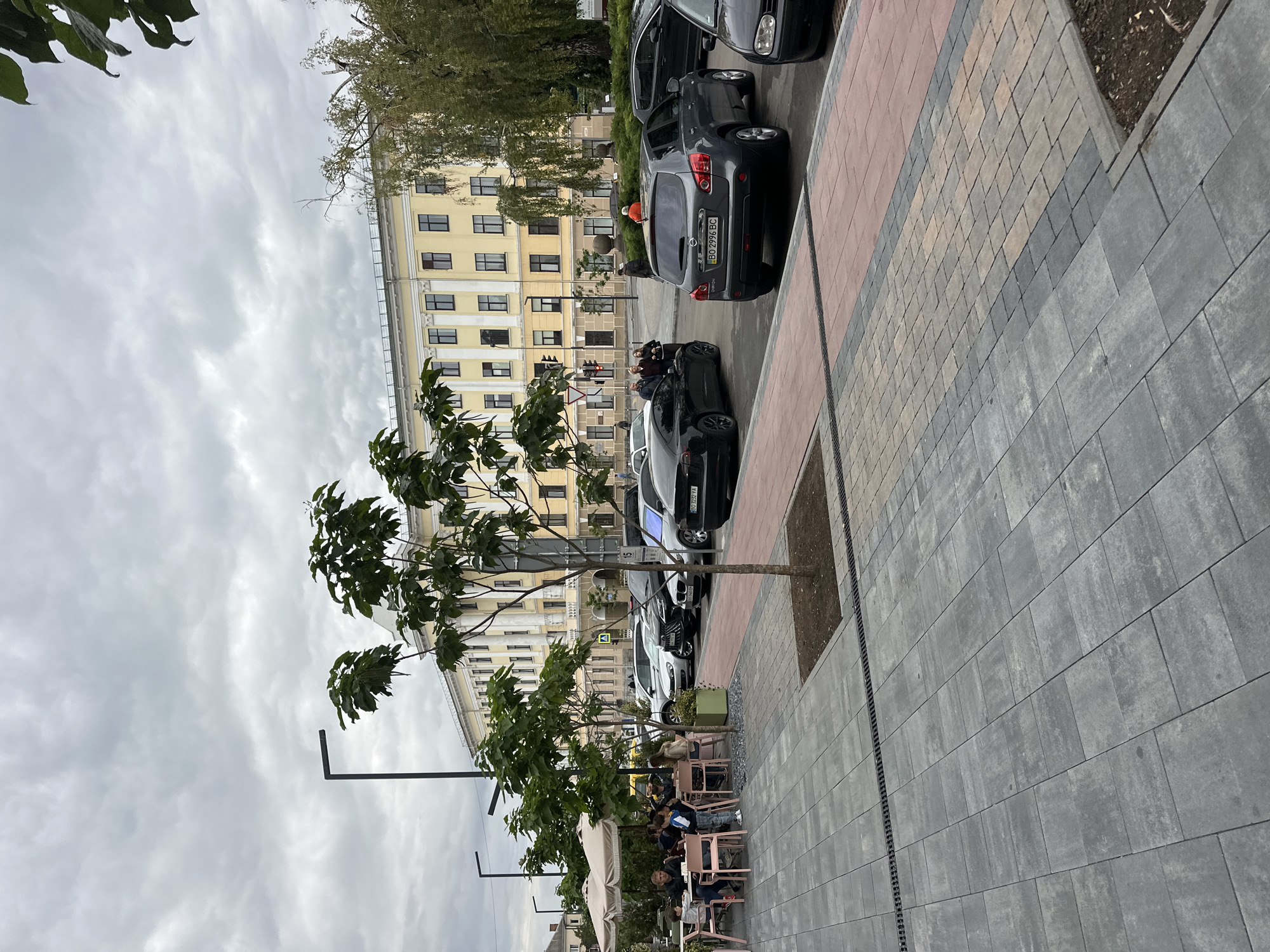 The image shows a view of the Ternopil City Council building, located at Teatralna Square in Ternopil, Ukraine. The building is a prominent, pale yellow structure with multiple windows and classical architectural elements. In the foreground, there is an outdoor seating area for a cafe with several tables and chairs, some occupied by people. A large umbrella provides shade over some of the tables. Several cars are parked on the street in front of the building, and a tree grows in the middle of the sidewalk. The pavement is made of grey and reddish-brown interlocking stones. The sky is overcast with grey clouds.