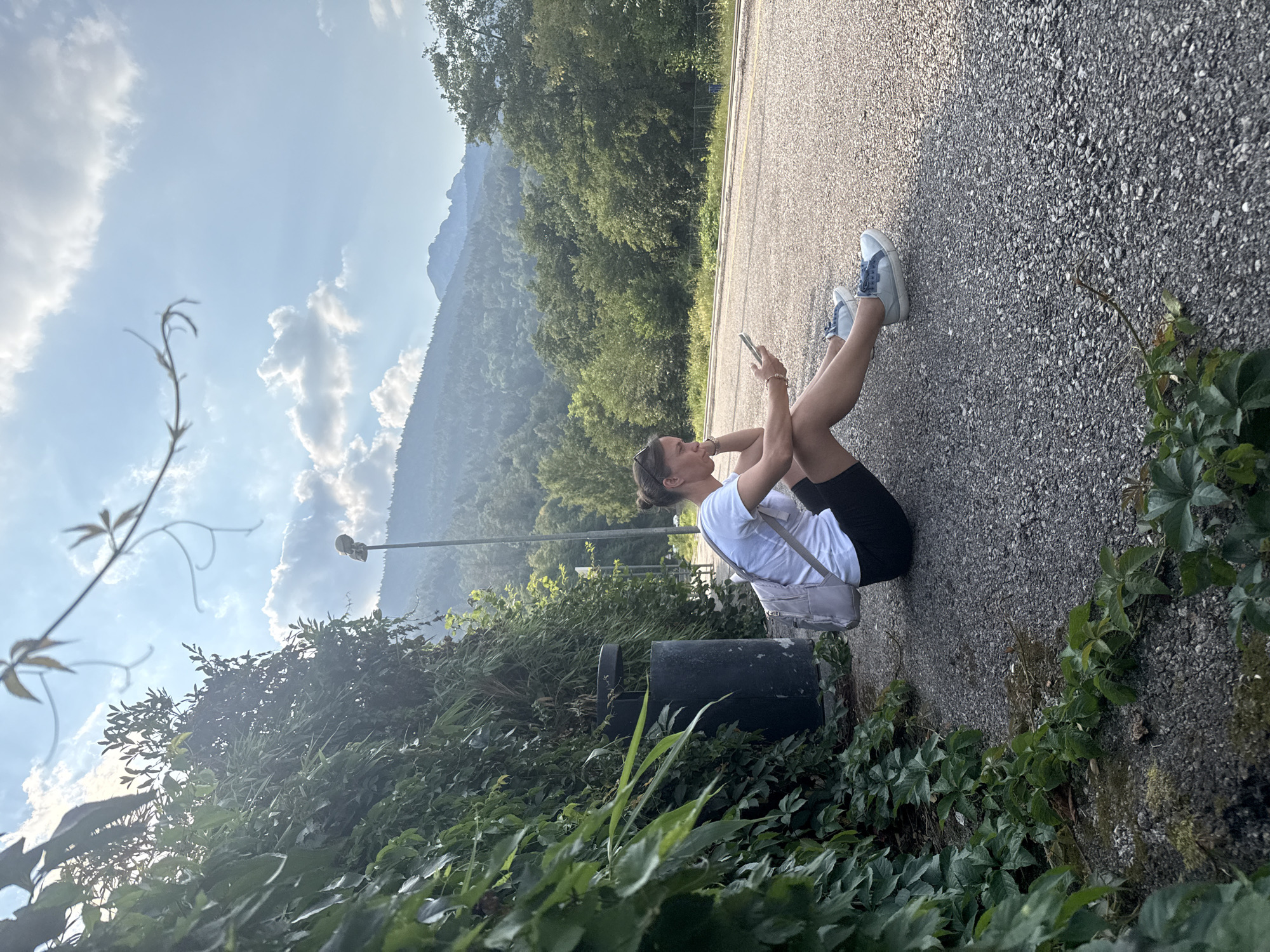 A woman is sitting on an asphalt road, holding a phone and looking at it. She is wearing a white t-shirt, black shorts, and blue and white sneakers. She has a light-colored bag slung across her chest. The woman has her legs bent and resting on the ground. In the background, there are trees and mountains. The sky is blue with some white clouds. The scene appears to be outdoors, possibly in a rural or mountainous area. The lighting suggests it is daytime, likely late afternoon or early evening, given the angle of the sun. There is no visible text in the image.