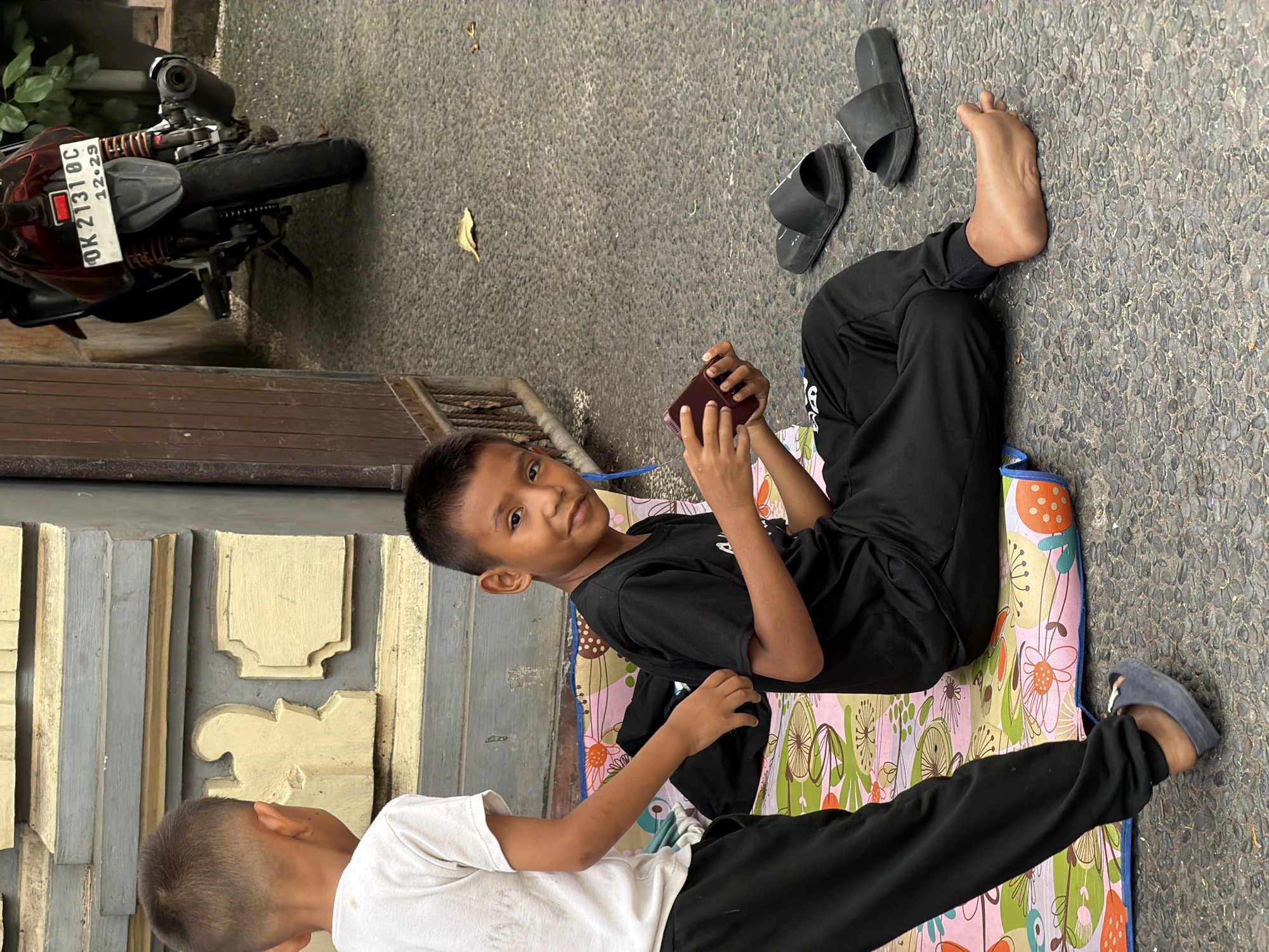 Two young boys are sitting on a colorful mat on a textured gray pavement. The boy on the right, wearing a black t-shirt and black pants, is holding a dark red mobile phone in his hands and looking towards the camera with a slight smile. He is barefoot. The boy on the left, wearing a white t-shirt and black pants, is partially visible from the side, facing away from the camera. He appears to be interacting with something on the mat. To the right of the boys, two black slide sandals are visible on the pavement. In the background, a motorcycle with a license plate reading "OK 2131 OC 12.29" is parked. Behind the motorcycle and the boys, there is a wall with decorative elements painted in light yellow and gray. A wooden gate is also visible to the left of the motorcycle. The environment appears to be outdoors in a street or courtyard setting.