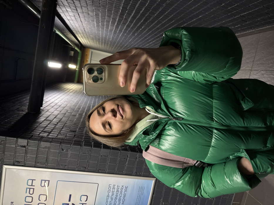 A woman in a shiny green puffer jacket takes a selfie indoors. She holds a smartphone horizontally, its screen facing the camera. The background consists of a tiled wall and what appears to be an underpass. A light fixture is visible in the distance. A sign with Cyrillic text is partially visible near the bottom of the frame.