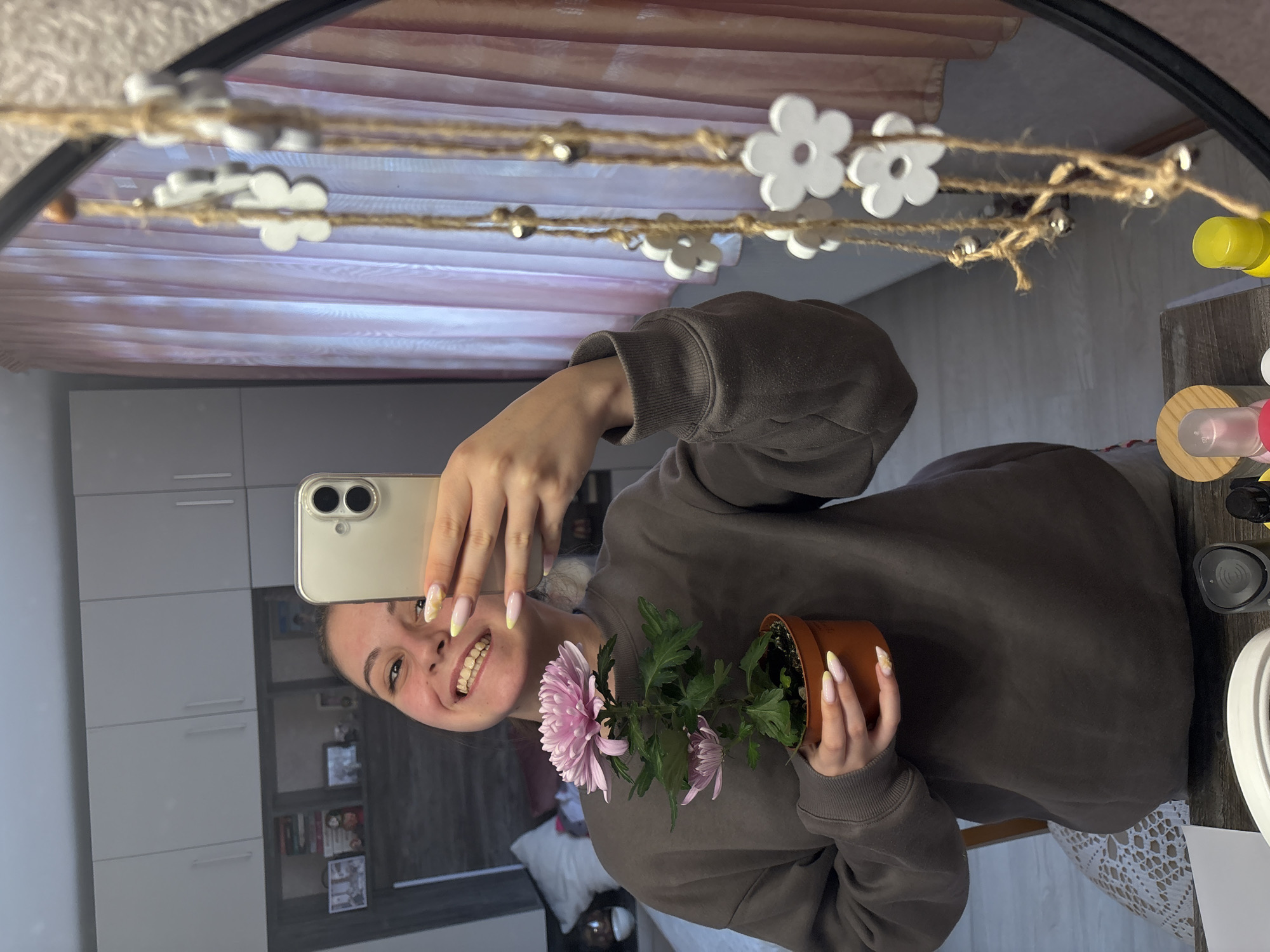 A young woman is taking a selfie in a mirror. She is holding a small potted plant with pink flowers. She is smiling and looking at the phone. The background shows a room with cabinets and a pink curtain. A string of white flower decorations hangs across the top of the mirror.
There is nothing in the photo to confirm this is Bulgarovo, Bulgaria.