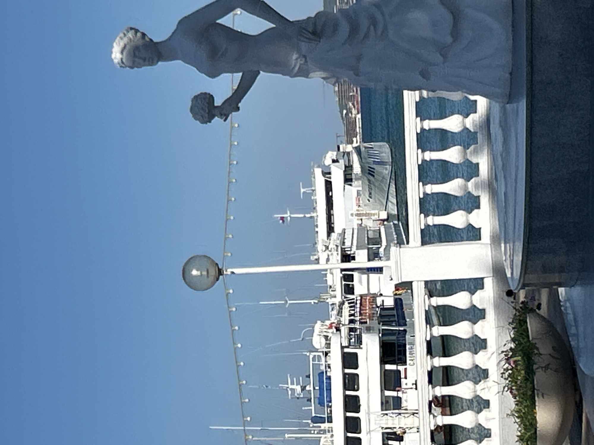 The image captures a serene waterfront scene in Gelendzhik, Russia, dominated by a clear blue sky and the tranquil blue sea. In the foreground, a classical statue of a woman holding a bouquet stands prominently, her form detailed in a light gray hue. She is positioned on a circular platform, with a decorative white balustrade separating her from the water. To the left of the statue, a large, textured planter holds lush green foliage.
Behind the balustrade, several white boats are docked, their sleek designs and multiple decks indicating they are likely passenger ferries or cruise vessels. The boats are adorned with windows and railings, reflecting the sunlight. A lamppost with a spherical light fixture stands to the left of the statue, casting a warm glow. In the background, the shoreline is dotted with buildings and lush greenery, suggesting the presence of a vibrant city. The overall atmosphere is one of peacefulness and beauty, with the calm sea and clear sky creating a picturesque setting.