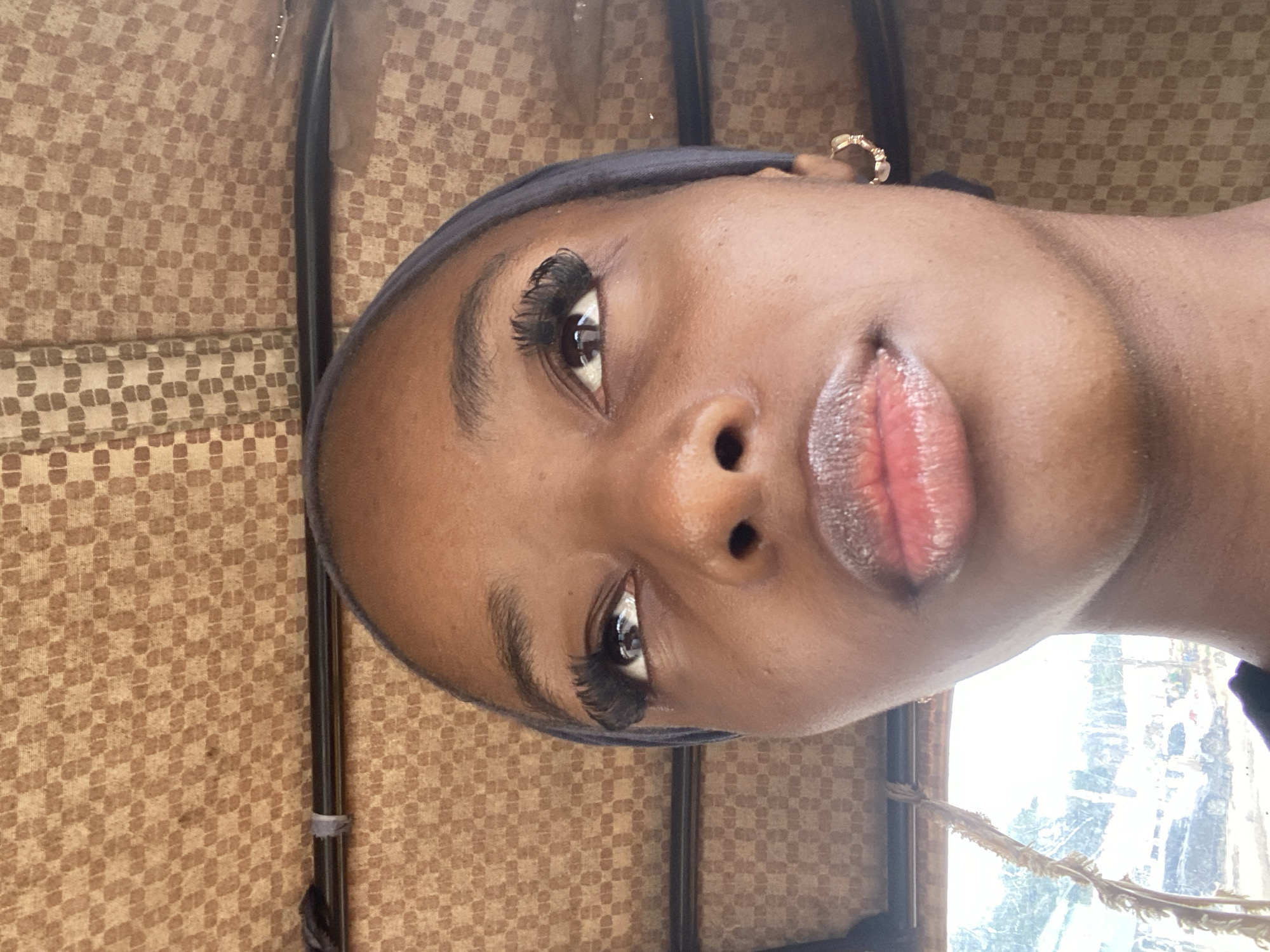 This is a close-up, slightly tilted photograph of a young Black woman's face. She is wearing a dark headscarf and has dramatic false eyelashes. Her lips are glossy and appear to be a reddish-pink color. She is wearing a small, gold hoop earring in her visible ear.
The background consists of a patterned fabric, likely upholstery or drapery, in shades of brown and tan, with a small section of a black pole visible. To the right of the woman's face, a bright, out-of-focus background suggests a window with daylight and possibly some greenery visible outside.
The lighting suggests it is daytime. There are no clear indications of a specific event or activity. The woman's expression is neutral, with her gaze directed slightly away from the camera. The overall impression is a selfie or a portrait taken in a casual setting, possibly indoors near a window. The location context provided is Jos, Nigeria. No visible text is present in the image.