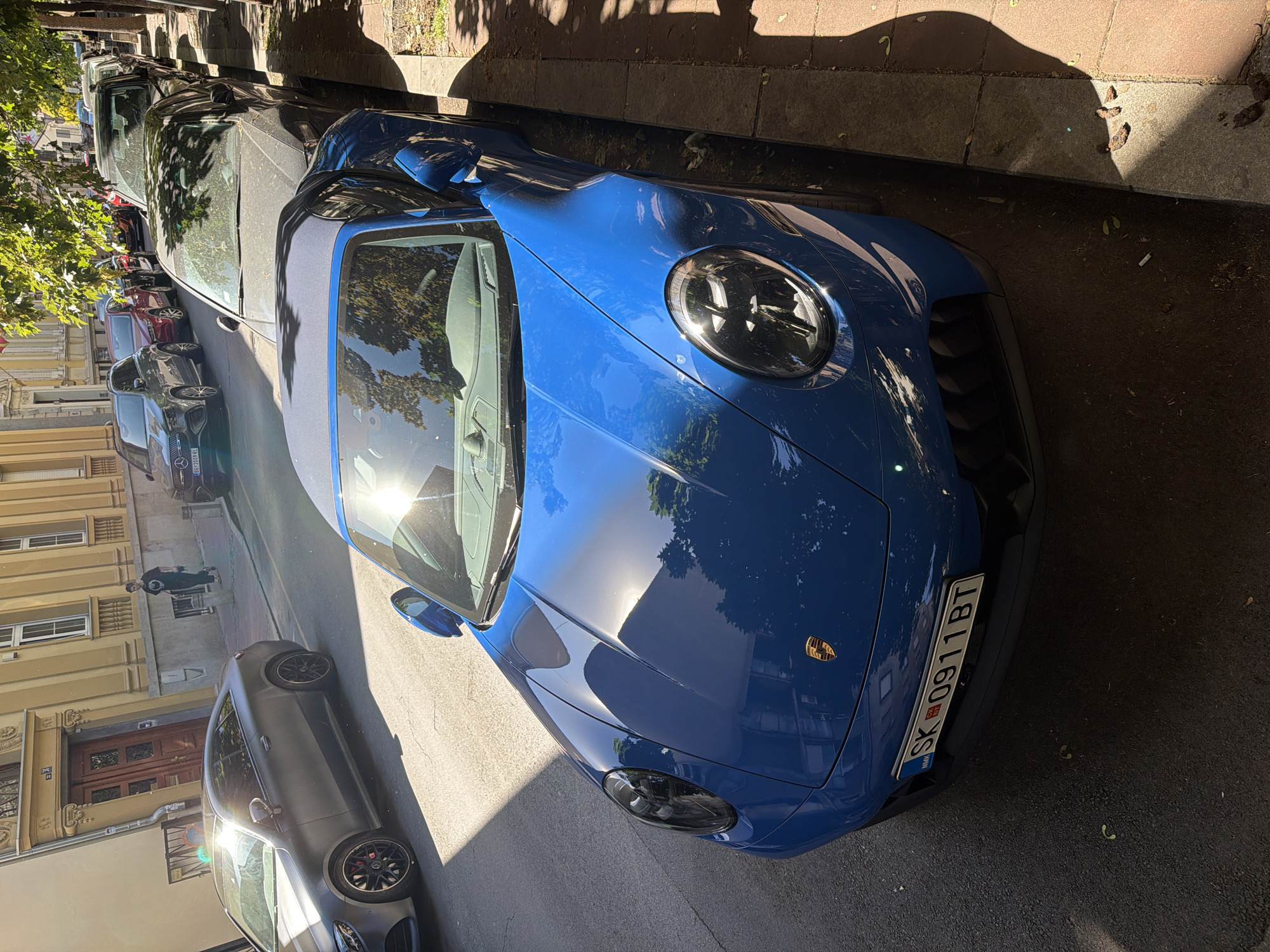 The image shows a blue Porsche parked on a street in Belgrade, Serbia. The license plate reads "SK 0911 BT," indicating it is a vehicle registered in Slovakia. The car is parked parallel to the curb, and several other vehicles are parked along the same side of the street. The lighting suggests it is daytime, and the angle of the sun creates reflections on the car's surface. The environment includes buildings on the opposite side of the street, trees casting shadows, and the typical street pavement. The focus is mainly on the Porsche, showcasing its design and color. A person is standing in the background, near a parked silver Mercedes-Benz. The scene is a common urban setting with parked cars in a residential or commercial street.
