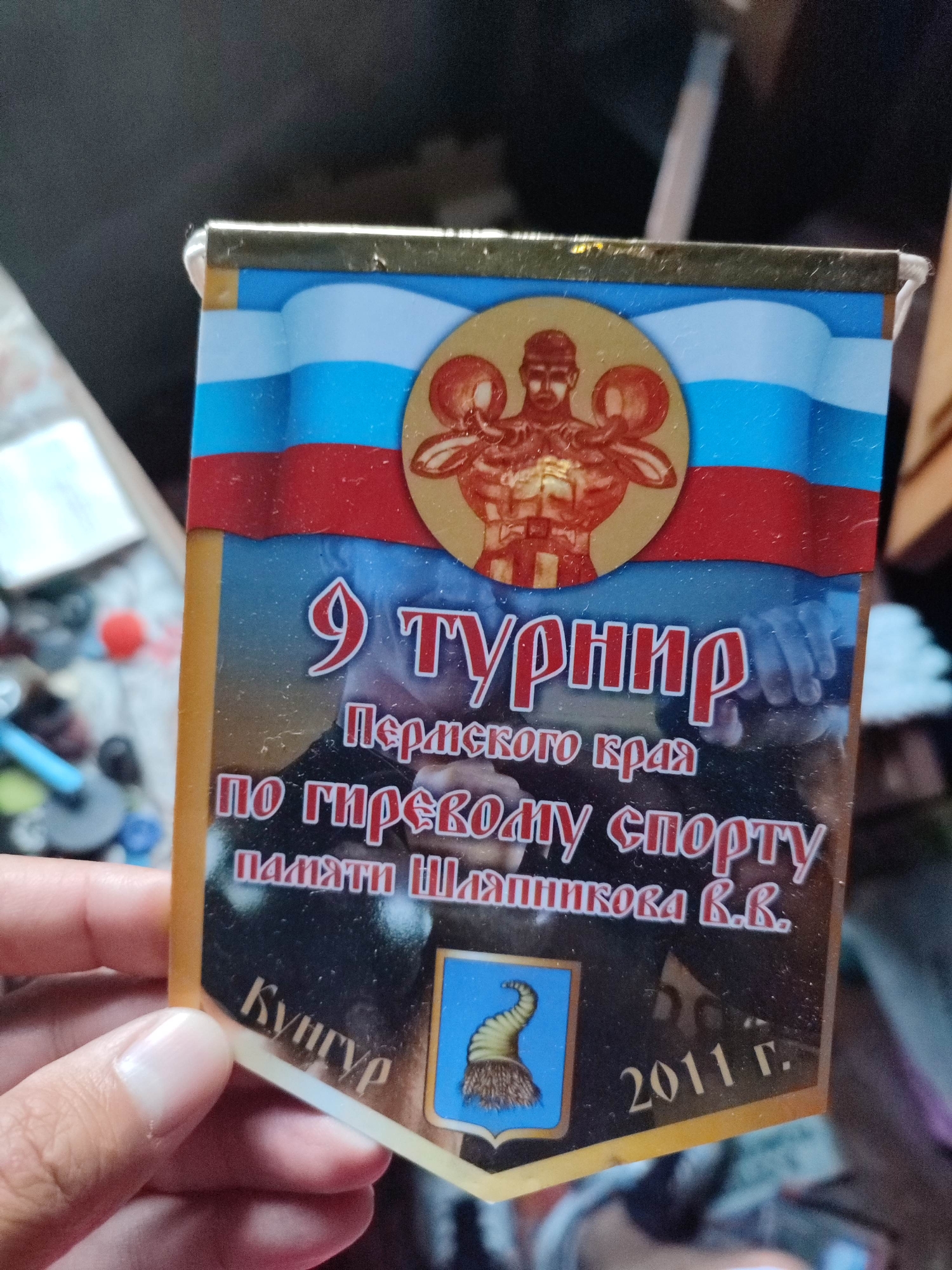 The image shows a close-up of a commemorative badge or medal. The badge is held in a person's hand, with a thumb visible on the left side. The badge features a design with the colors of the Russian flag at the top, red, white, and blue stripes. In the center, a circular emblem depicts a muscular, bare-chested male figure from the chest up, holding two large, round weights over his shoulders. This emblem is surrounded by a gold border.
Below the emblem, text is prominently displayed in large, stylized red lettering. The first line reads "9 ТУРНИР" (9th Tournament). The second line, in smaller white text, says "Пермского края" (of Perm Krai). The third line, again in large white text, states "ПО ГИРЕВОМУ СПОРТУ" (for Kettlebell Sport). The fourth line, in smaller white text, reads "ПАМЯТИ ШАПНИКОВА В.В." (in memory of Shapnikov V.V.).
On the lower left of the badge, the word "Кунгур" (Kungur) is written in white, curved lettering. To the right of this, a small blue shield with a golden, fur-like tuft or horn shape is visible. On the lower right side of the badge, the year "2011 г." (2011) is printed in white.
The badge appears to be made of metal and possibly enamel or printed material. The overall impression is that of an award or souvenir from a sporting event. The background is blurry but suggests an indoor setting with some objects and light sources. The lighting in the image is somewhat dim, and there are some dust specks visible on the badge, indicating it might not be a professional studio shot but rather a casual photograph. The event commemorated is the 9th Kettlebell Sport Tournament of Perm Krai, held in memory of V.V. Shapnikov. The location cues, "Perm Krai" and "Kungur," place the event within Russia.