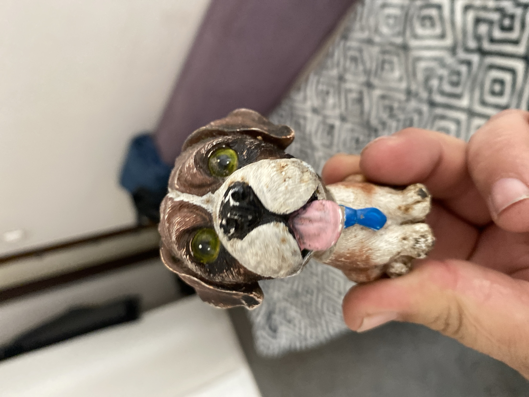 A close-up, eye-level shot shows a person's hand holding a small, detailed figurine of a dog. The dog, which appears to be a Saint Bernard breed, has brown and white fur, green eyes, and its pink tongue is sticking out. It's wearing a small blue tie. The background is blurred, showing what appears to be an indoor setting with a patterned surface and furniture. The image does not contain information to determine its location in Szczecin, Poland.