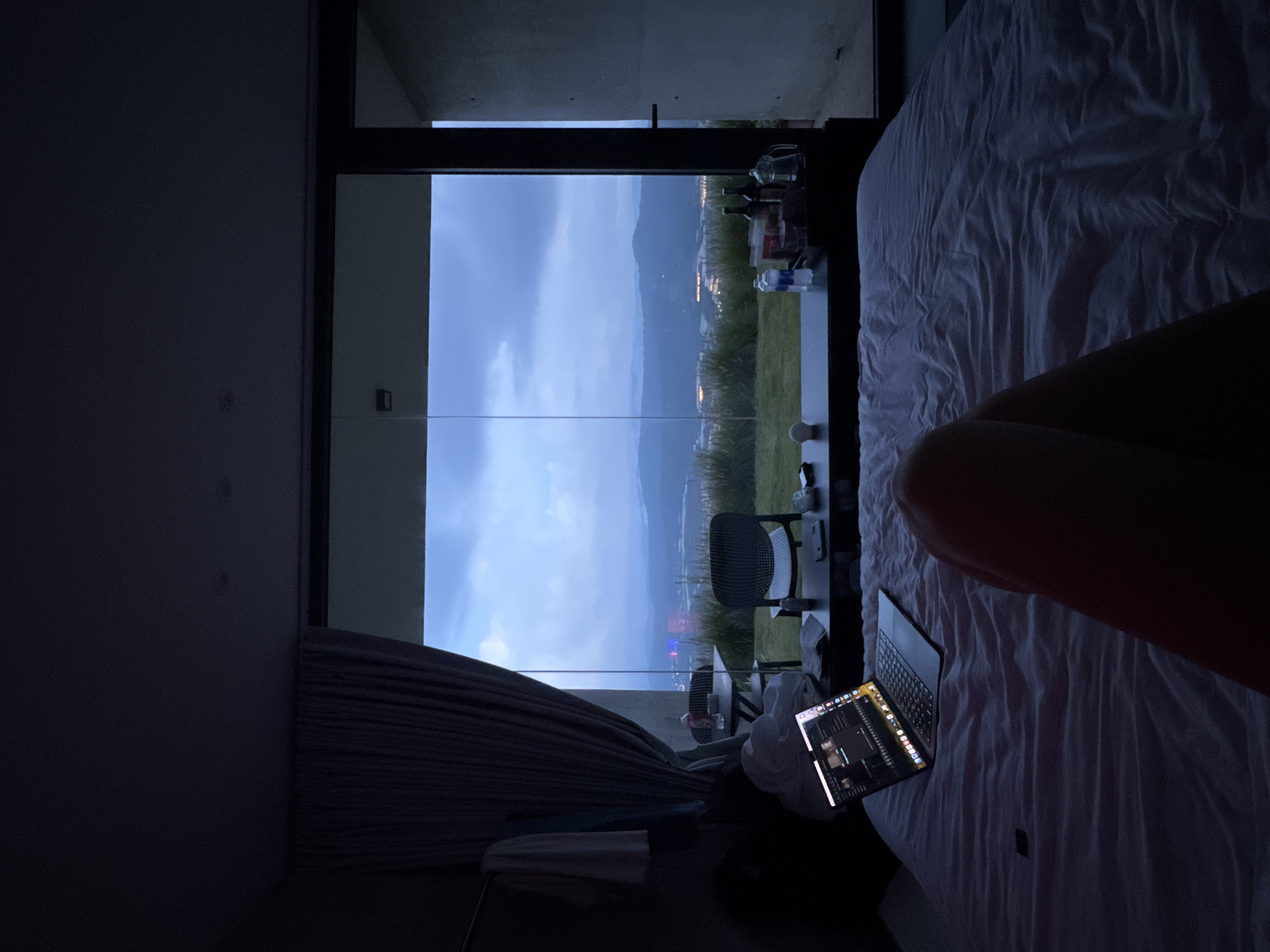 The image shows a dimly lit bedroom with a large window offering a panoramic view of a city at dusk. The room features a bed in the foreground, covered with a crumpled white duvet. A laptop is open on the bed, displaying a video editing timeline. To the left of the bed, a curtain hangs partially drawn. Outside the window, a landscape unfolds with rolling hills, vegetation, and the illuminated city lights. Two chairs and a small table are visible on a grassy area beyond the window, and on the table, there are bottles and a pitcher. The sky is overcast with hues of blue and grey. The scene appears to be set in a hotel room or a modern residence in a hilly area overlooking the city of Da Loc, Vietnam.